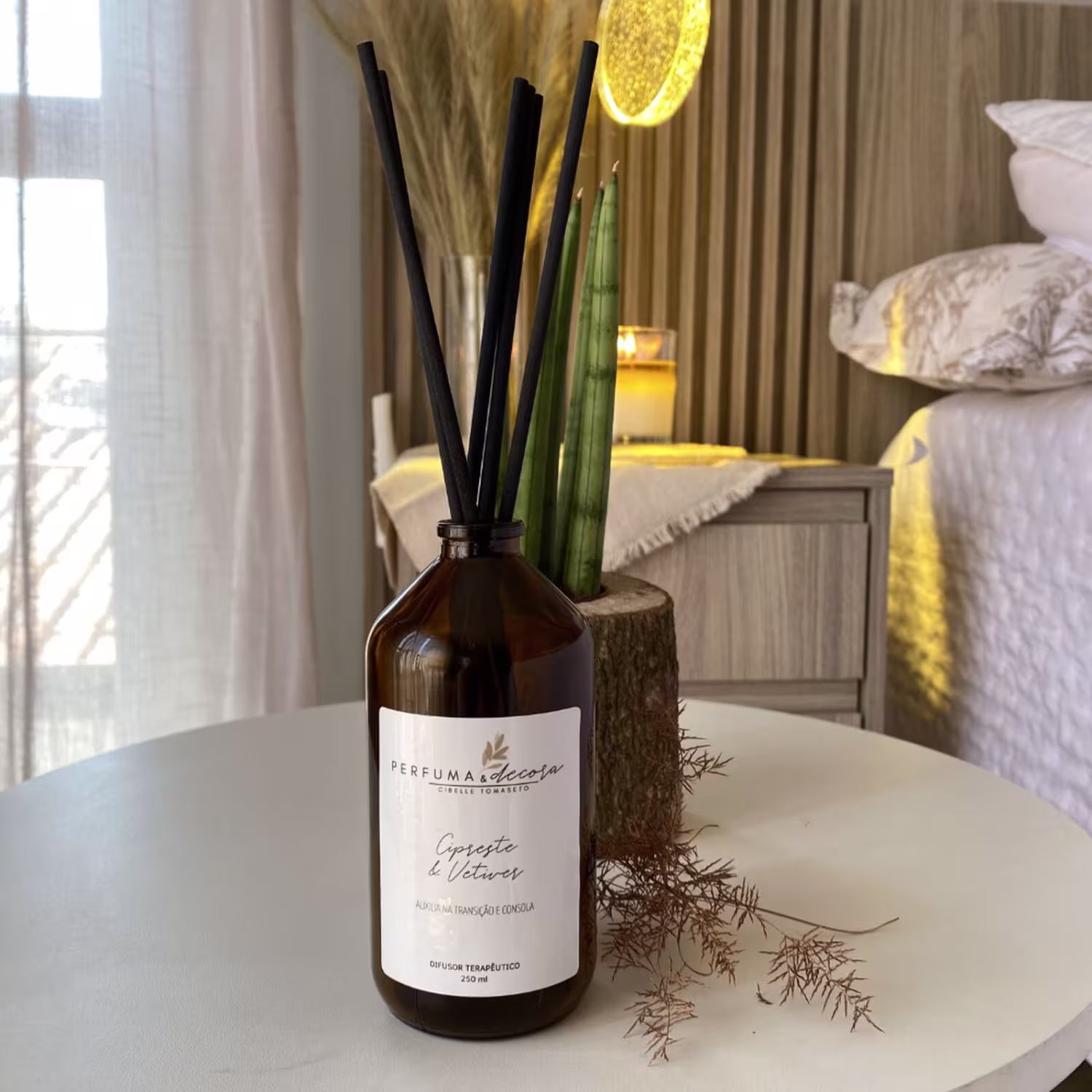 Difusor de ambiente em frasco de vidro âmbar. Varetas de difusor pretas e verdes inseridas no frasco. Vela amarela acesa e decoração de palha ao lado. Cores neutras e terrosas no ambiente, com almofadas brancas e detalhes amarelos.