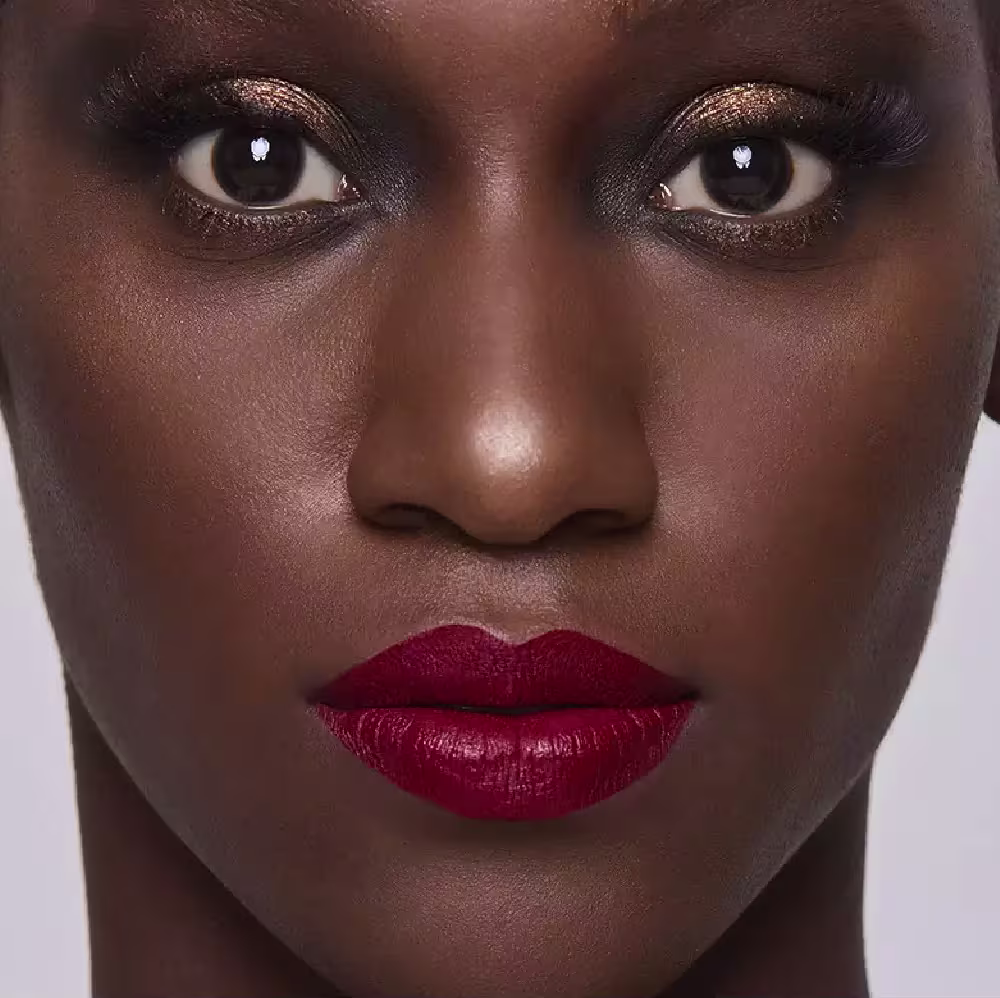 Esta imagem mostra uma mulher negra com maquiagem elaborada. A pele tem um lindo tom marrom vibrante e brilhante. Os lábios foram maquiados com um batom de cor vermelha, realçando a boca bem delineada e cheia.