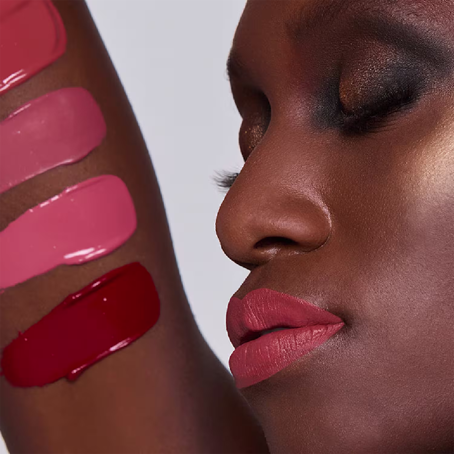 Esta imagem mostra uma mulher negra com maquiagem elaborada. A pele tem lindo tom marrom vibrante e brilhante. Os lábios estão com batom de cor rosa avermelhado envelhecido, realçando a boca bem delineada e cheia. Com amostras das cores em seu braço.