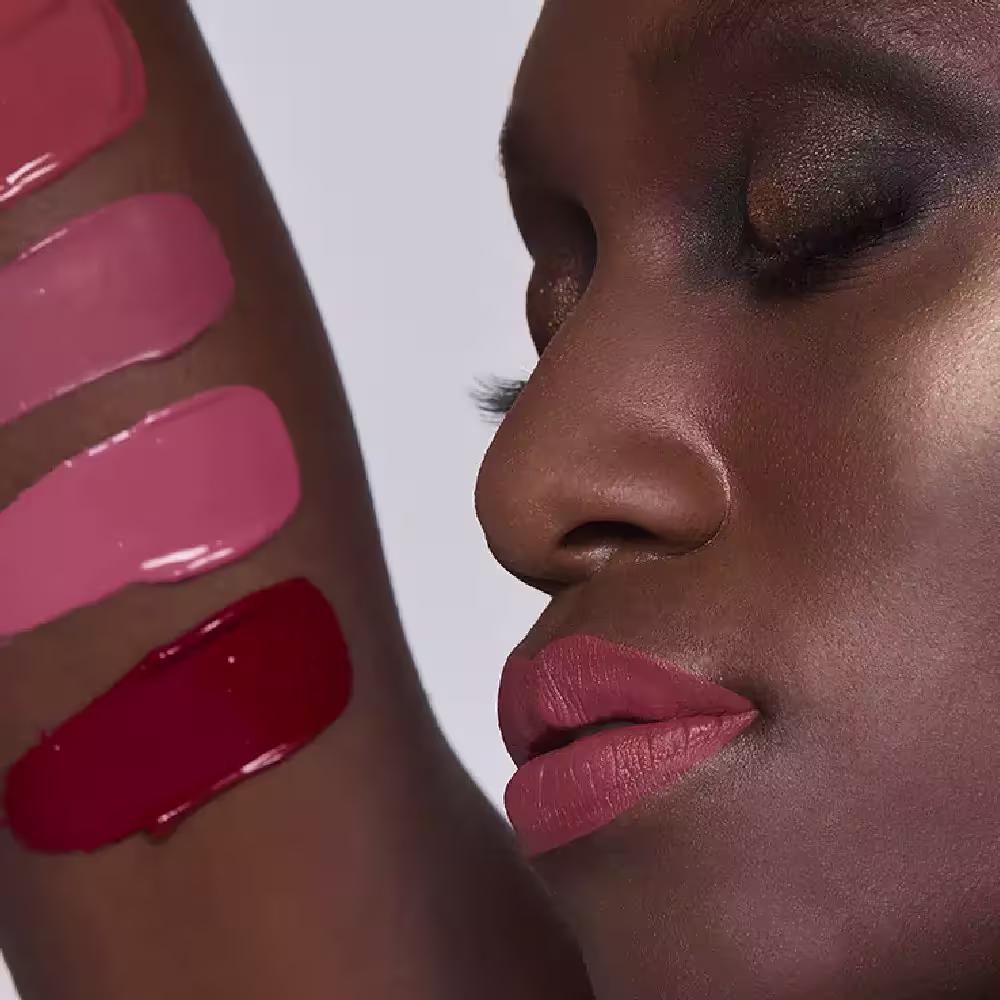 Esta imagem mostra uma mulher negra com maquiagem elaborada. A pele tem lindo tom marrom vibrante e brilhante. Os lábios estão com batom de cor rosa avermelhado envelhecido, realçando a boca bem delineada e cheia. Com amostras das cores em seu braço.
