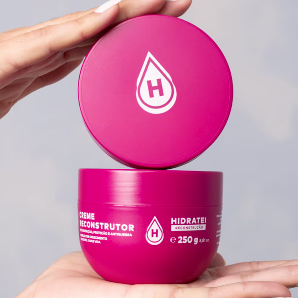 Embalagem 250g de plástico rosa vibrante redondo. A tampa apresenta o logotipo da marca, uma gota estilizada com a letra "H" no centro. As mãos seguram o pote e a tampa, exibindo o produto de forma clara.