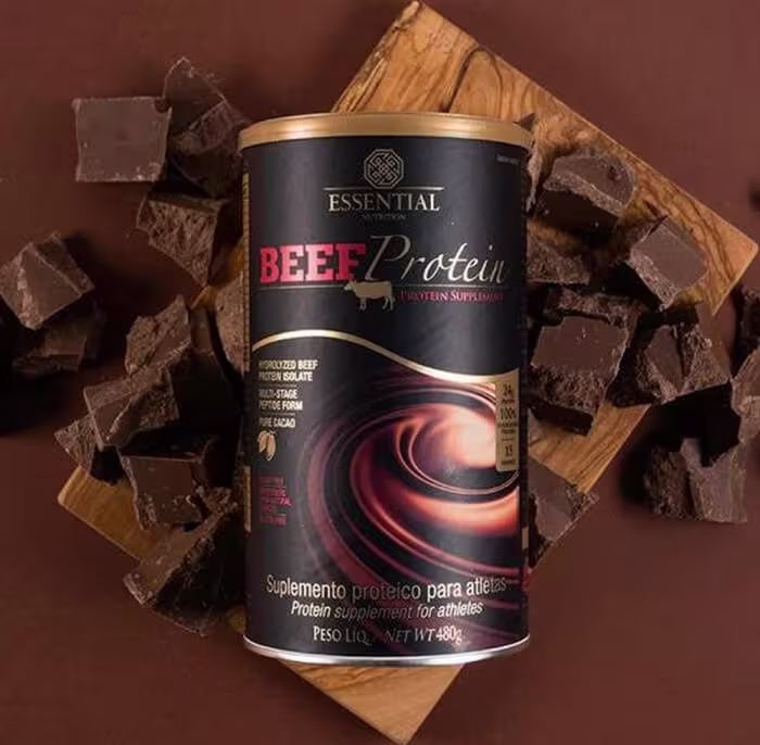 A imagem mostra uma lata de suplemento com fundo preto em meio a pedaços de chocolate.
