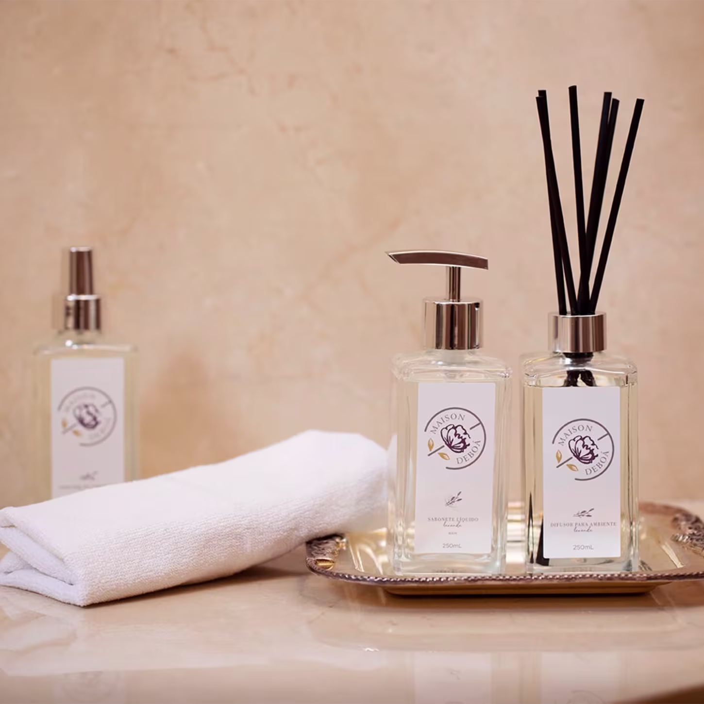 Produtos de fragrância e cuidados corporais da marca Maison Deboá. 3 frascos de vidro transparente com tampas douradas.. Um frasco é um pulverizador, outro tem um dosador de bomba e o terceiro contém difusores de aromas. Também há uma toalha branca