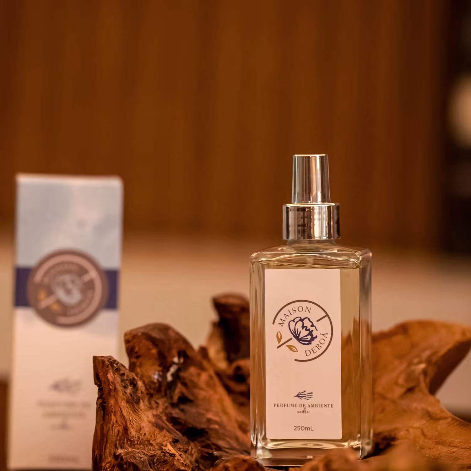 Perfume de ambiente. Ao lado, uma caixa branca com o mesmo logotipo azul. Folhas secas marrons e uma estrutura de madeira ao redor do frasco criam um cenário rústico.