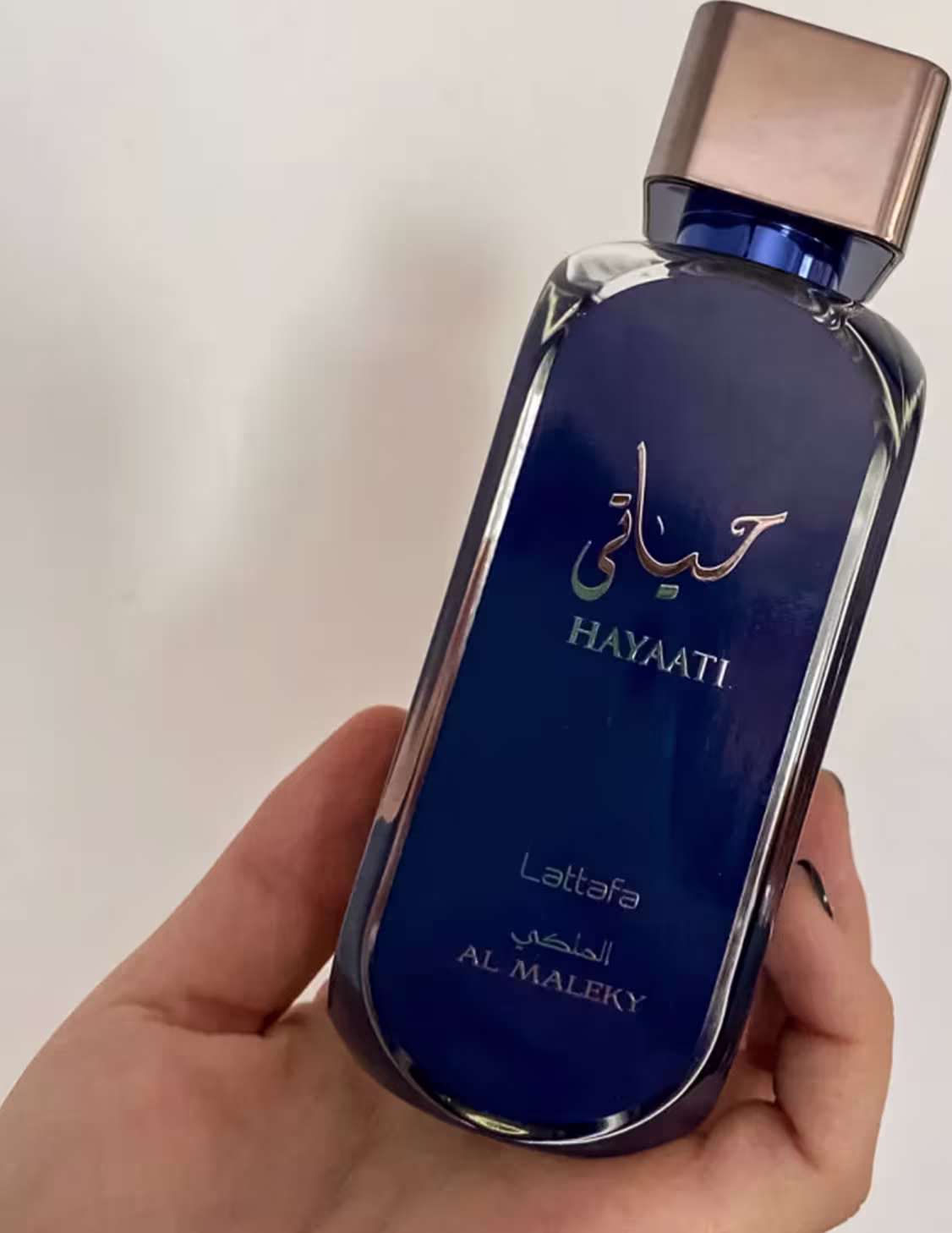 A imagem mostra o perfume sendo segurado por uma mão feminina.