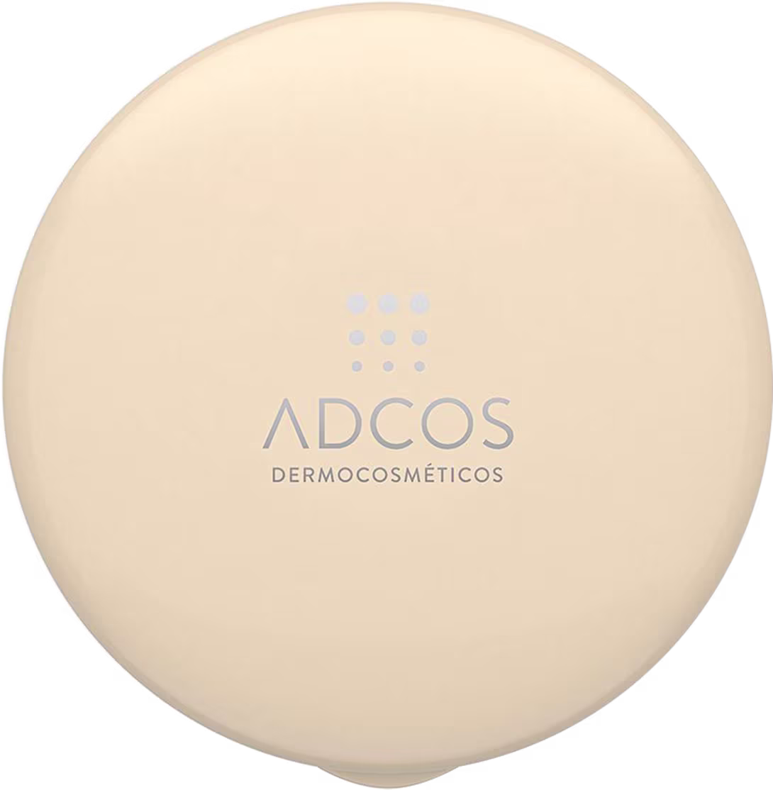 Protetor solar em pó em embalagem redonda e achatada. A embalagem é de cor bege claro. No centro, há o logotipo da marca Adcos Dermocosméticos impresso em letras cinza claro. O produto apresenta uma aparência simples e minimalista.