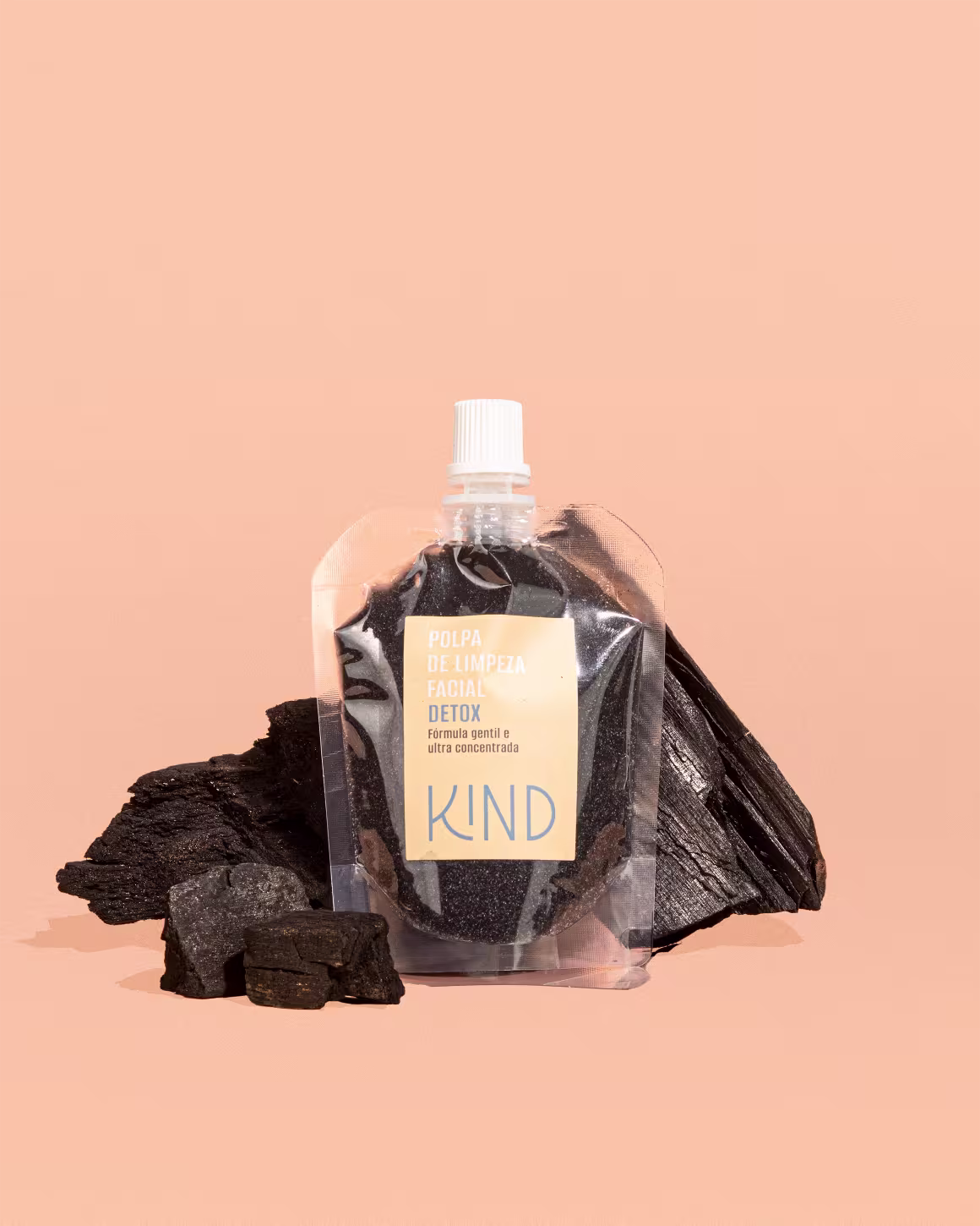 A imagem apresenta a embalagem da polpa de limpeza facial em um fundo laranja claro, com alguns pedaços de carvão atrás da embalagem.