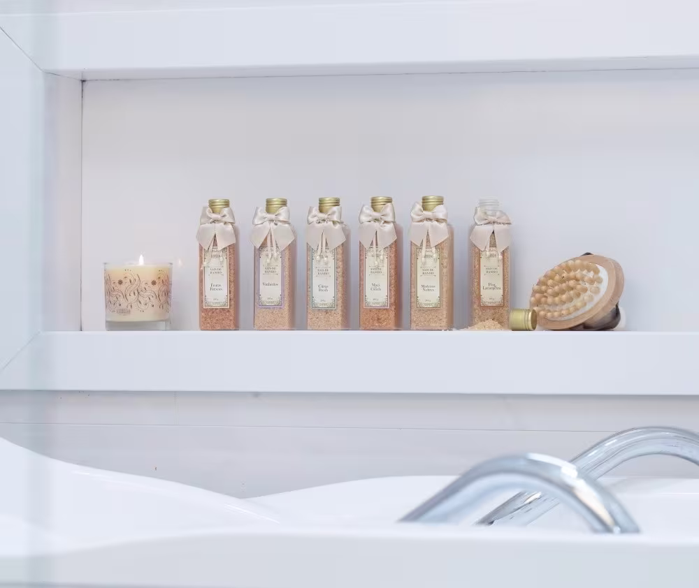 Seis frascos de vidro com tampas douradas e laços de tecido, contendo produtos da marca Empório Essenza Serra Gaúcha, exibidos em uma prateleira branca junto a uma vela decorativa e uma escova de massagem.