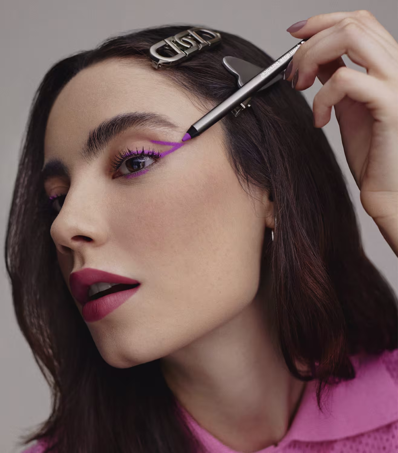 A imagem mostra uma modelo usando maquiagem de olhos em tons de roxo vibrante. Ela tem sobrancelhas bem delineadas e batom vermelho escuro nos lábios. Seu cabelo castanho escuro está penteado para trás, permitindo ver claramente seu rosto.