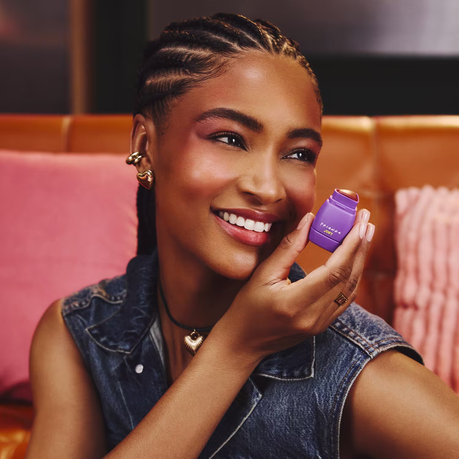 A pessoa sorri enquanto segura um item de maquiagem roxo em um ambiente com decoração rosa e laranja. Ela usa jaqueta jeans e acessórios dourados. O cenário sugere um ambiente informal e acolhedor, provavelmente relacionado a um produto de beleza.