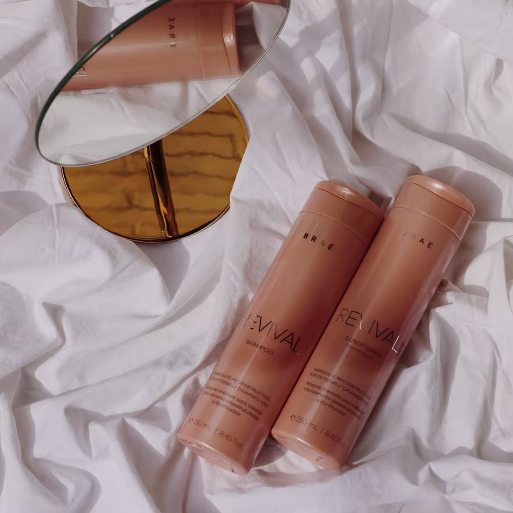 A imagem mostra shampoo e condicionador Revival da Braé. Eles estão deitados e ao lado dos frascos, há um espelho de maquiagem redondo com moldura dourada. Os itens estão dispostos sobre um tecido branco amarrotado.