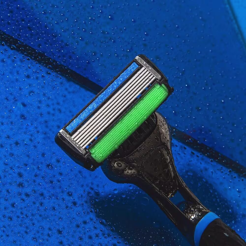 A imagem mostra um aparelho de barbear descartável. Possui um corpo plástico preto com detalhes em azul. A lâmina de barbear tem uma faixa azul e outra verde, com ranhuras metálicas expostas.