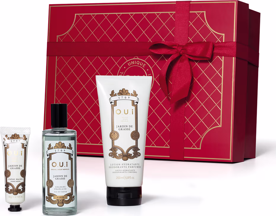A imagem mostra um conjunto de produtos O.U.i em frente a uma bela caixa de presente vermelha. Ela traz um frasco com eau de parfum e dois tubos brancos com tampa preta, um menor com hidratante para as mãos e outro maior com hidratante para o corpo.