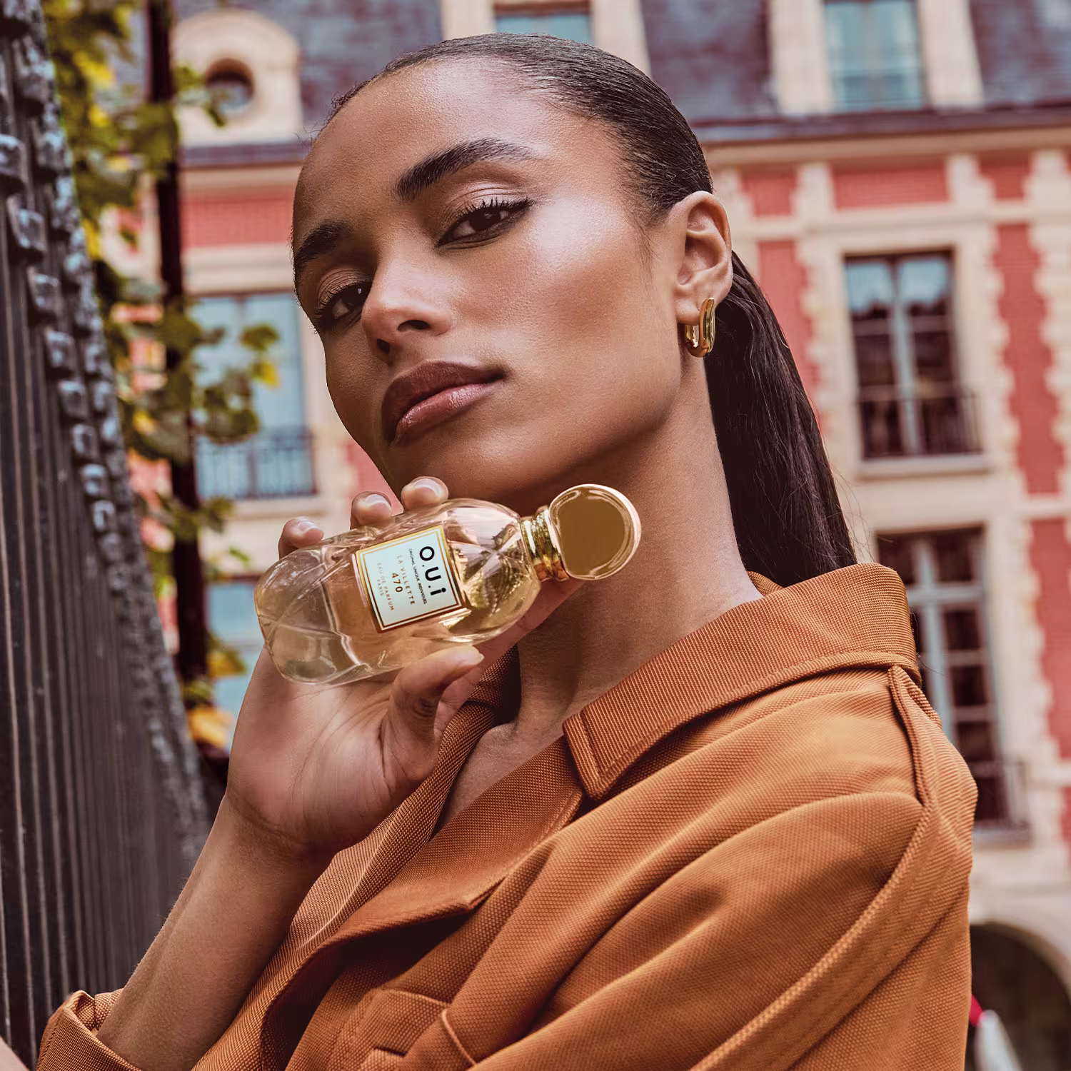 A mulher segura um frasco de perfume da marca Oui em uma rua urbana com edifícios históricos ao fundo, vestindo um casaco laranja, sugerindo um ambiente sofisticado e elegante para o uso do produto.