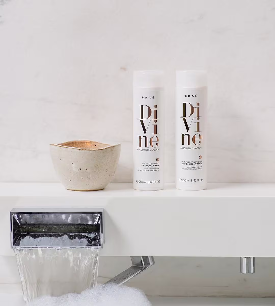 Dois frascos de shampoo e condicionador da linha Divine da marca Braé. Os frascos são cilíndricos, brancos e apresentam o logotipo e nome "Divine" em letras marrons e pretas.
