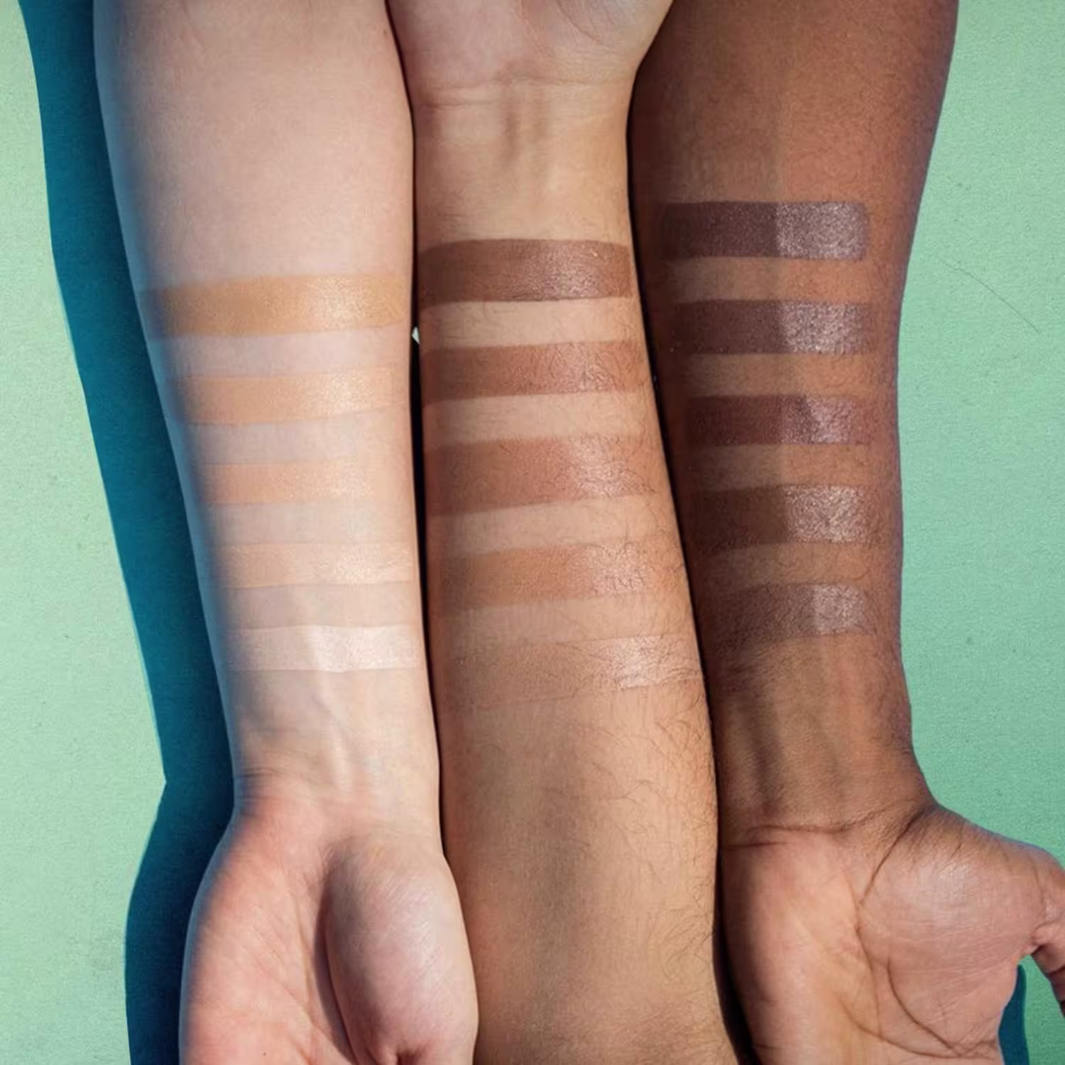 A imagem mostra três antebraços com tons de peles diferentes, num fundo verde claro. Na pele, amostras das 15 tonalidades dos produtos, 5 cores em cada antebraço.
