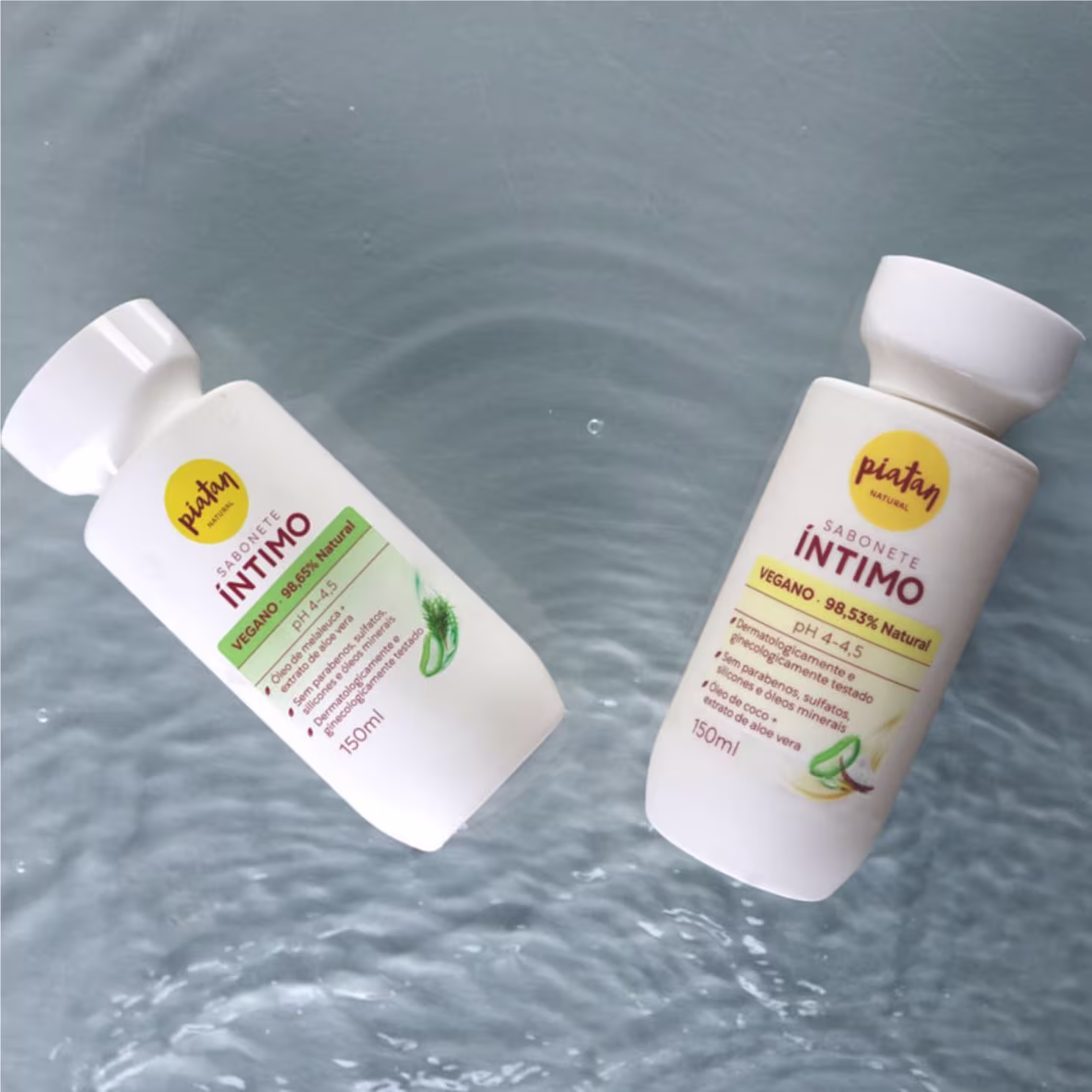 Dois frascos de sabonete íntimo vegano da marca Piatan. Um frasco maior de 150ml com rótulo amarelo e texto "Íntimo Vegano 98,53% Natural" e um frasco menor com rótulo verde e texto "Sabonete Íntimo Vegano". Os frascos estão em uma superfície líquida.