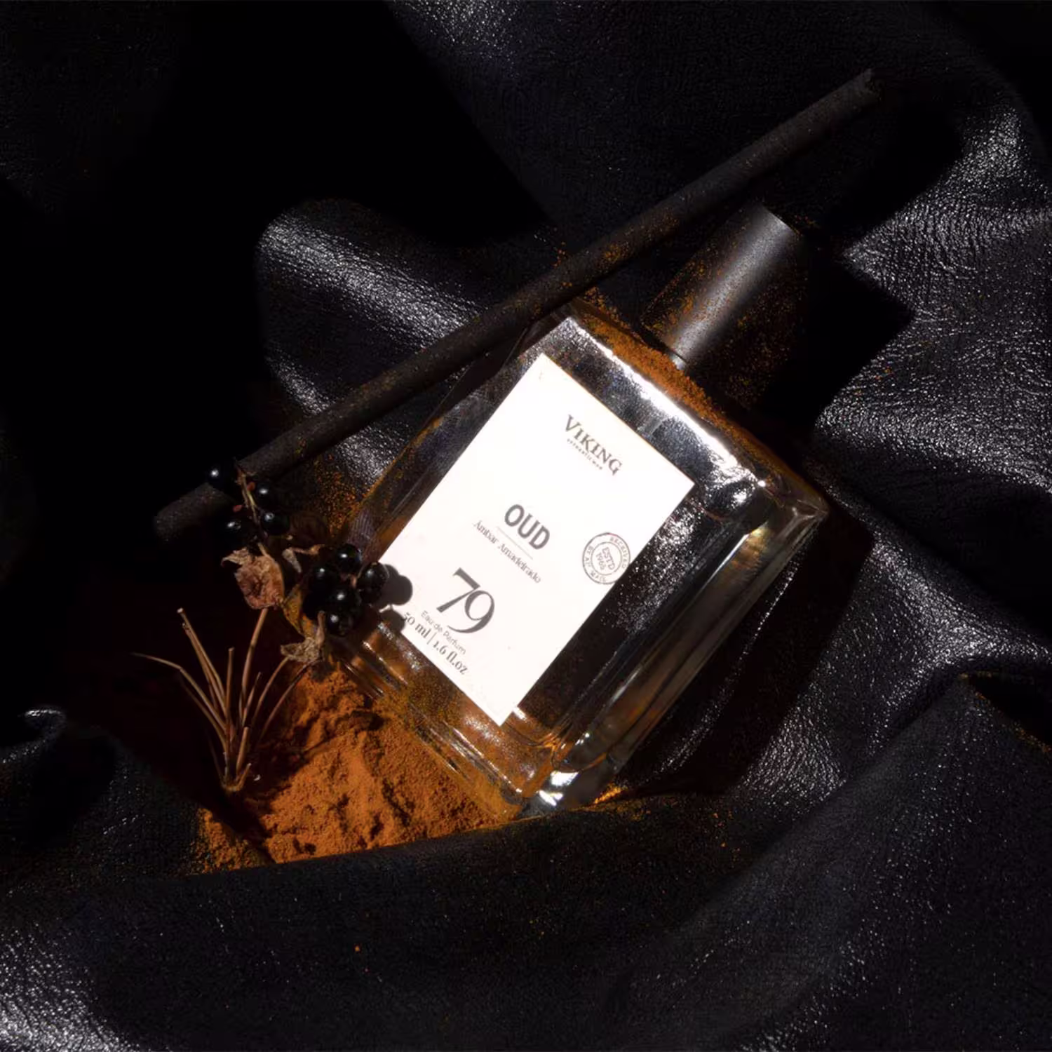 Perfume masculino em frasco retangular de vidro âmbar. Rótulo branco com o nome "Oud 79" e o logotipo Viking. Sobre um fundo preto texturizado, acompanhado de galhos secos, grãos de café e especiarias em pó marrom.