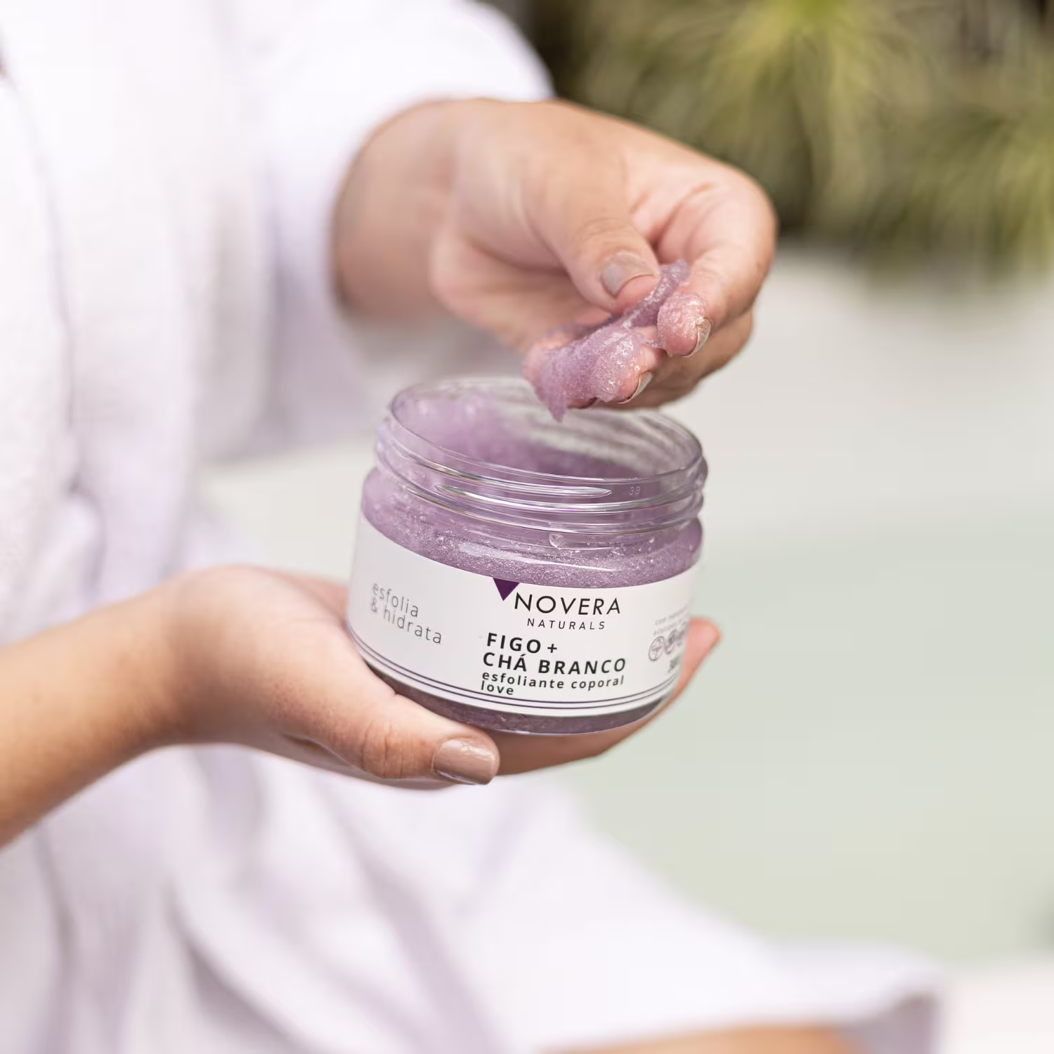 A imagem mostra duas mãos segurando um pequeno pote de vidro transparente contendo um produto corporal esfoliante natural. O produto tem uma cor roxa clara e parece ter uma textura cremosa.
