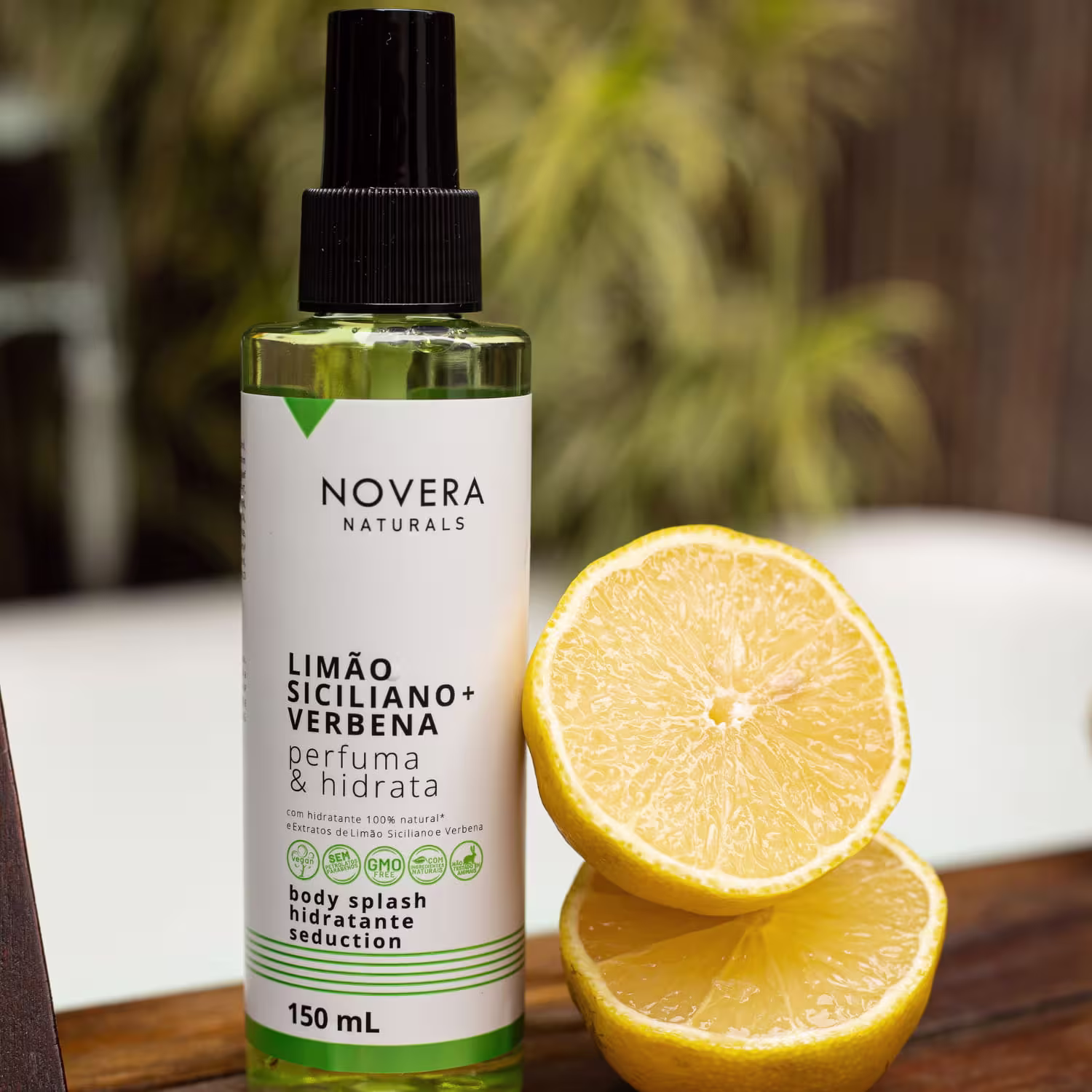 A imagem mostra body splash Novera Naturals com fragrância de limão siciliano e verbena. O frasco é transparente, tem 150ml de capacidade e está acompanhado de uma fatia de limão aberta ao meio, evidenciando os detalhes suculentos do cítrico.