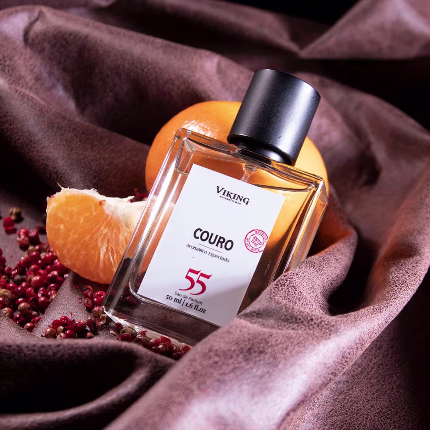 Perfume Viking em frasco retangular transparente. Embalagem com rótulo branco exibindo o nome "Couro 55" e o logotipo Viking. Tampa preta fosca. Apresentado sobre tecido marrom com pedaços de couro cru e grãos vermelhos espalhados ao redor.