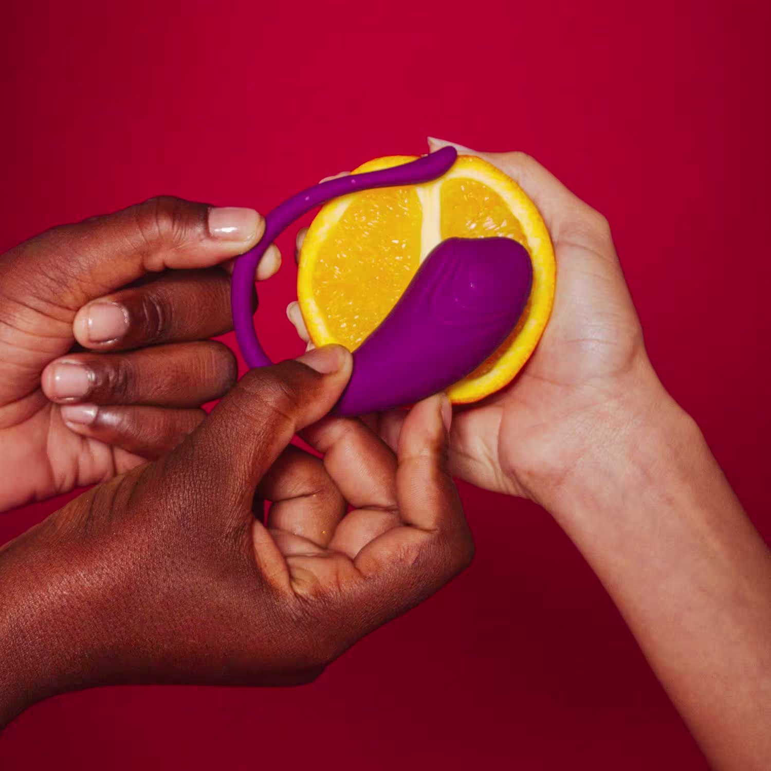 Mãos segurando uma laranja cortada ao meio e um vibrador curvo de cor roxa, em um fundo vermelho vibrante.