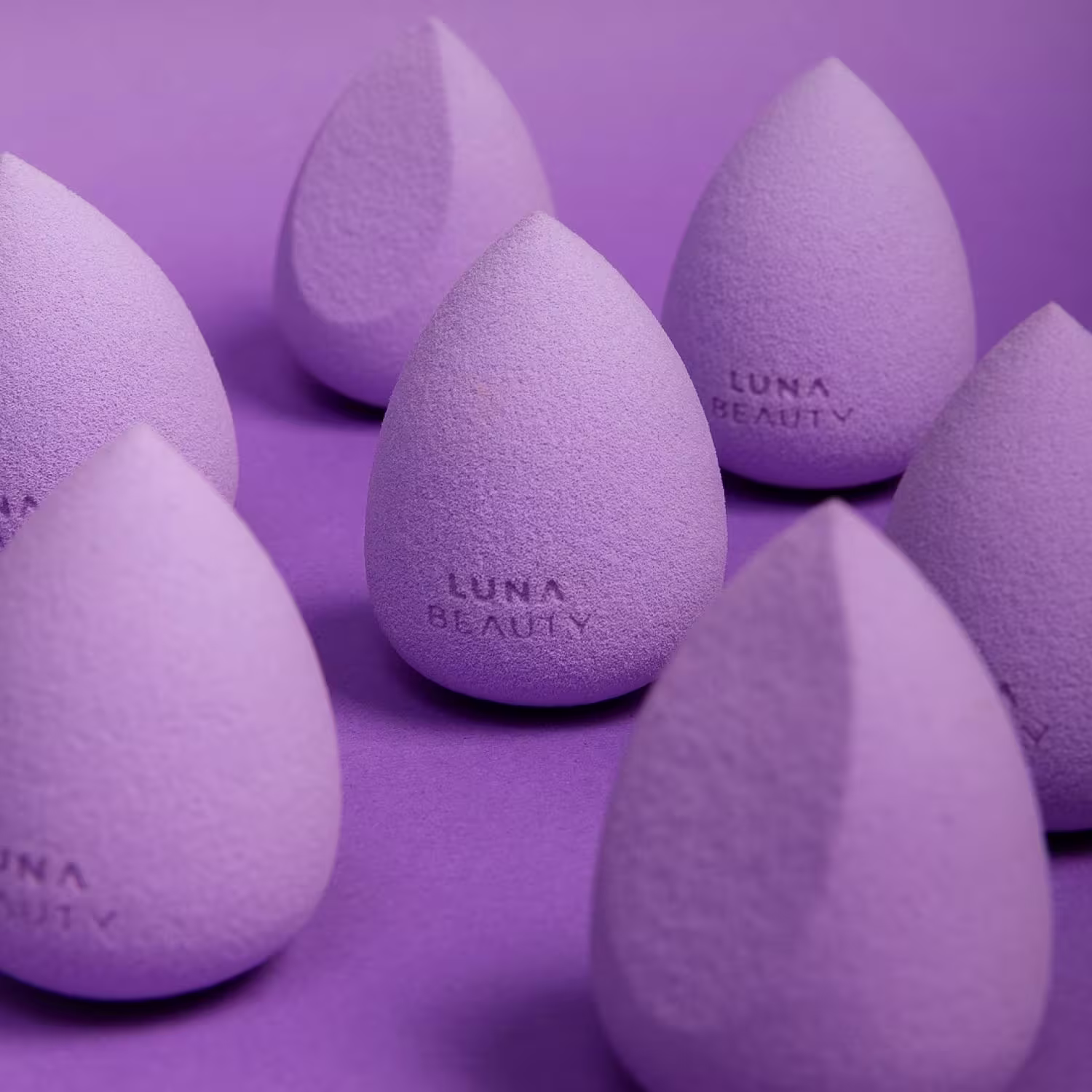 Esponjas de maquiagem na forma de ovos. As esponjas são de cor lilás e têm a inscrição "Luna Beauty" gravada em algumas delas. Apresentam uma textura aveludada e estão dispostas em um arranjo agrupado sobre um fundo monocromático na mesma tonalidade lilás
