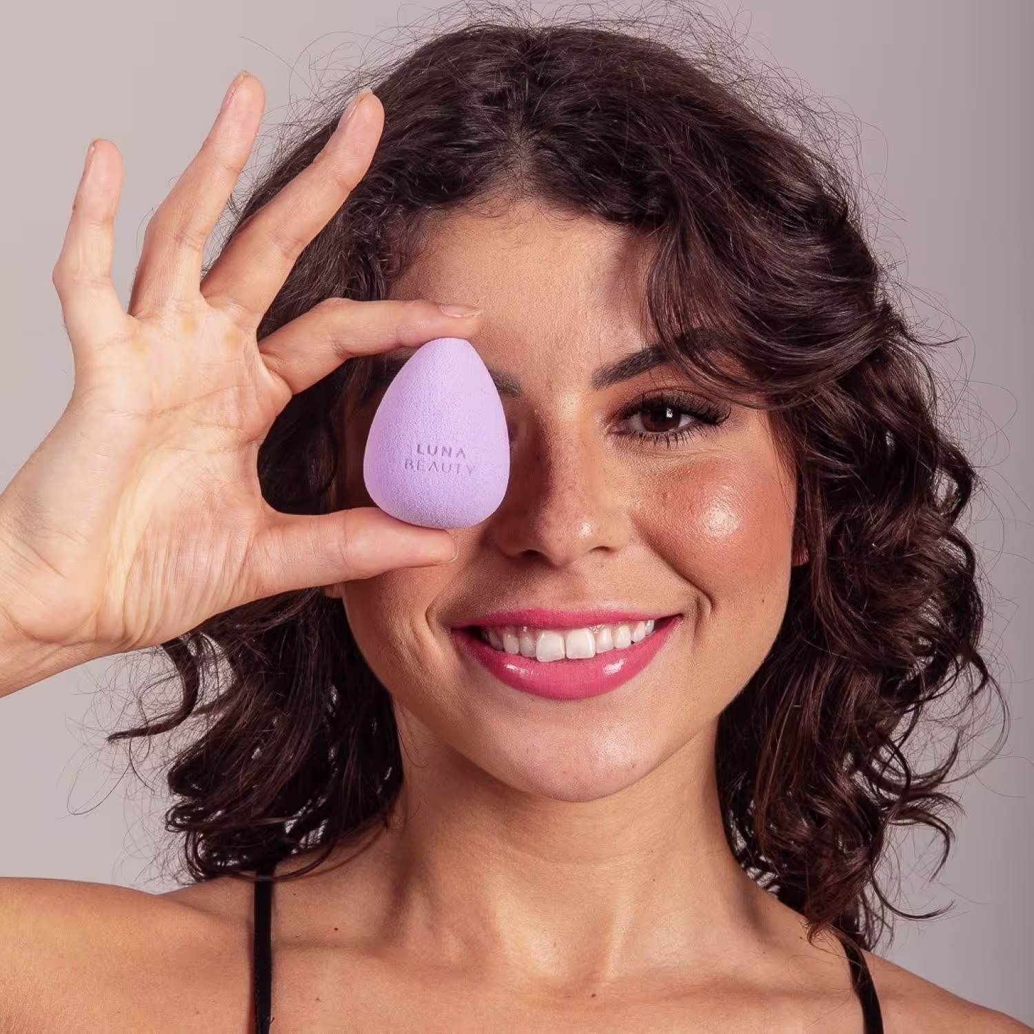 Esponja de maquiagem na forma de um ovo de cor lilás. A esponja está sendo segurada próxima ao rosto sorridente de uma mulher de cabelos escuros e ondulados. A imagem tem um fundo neutro e iluminado que destaca o produto e o rosto da modelo.
