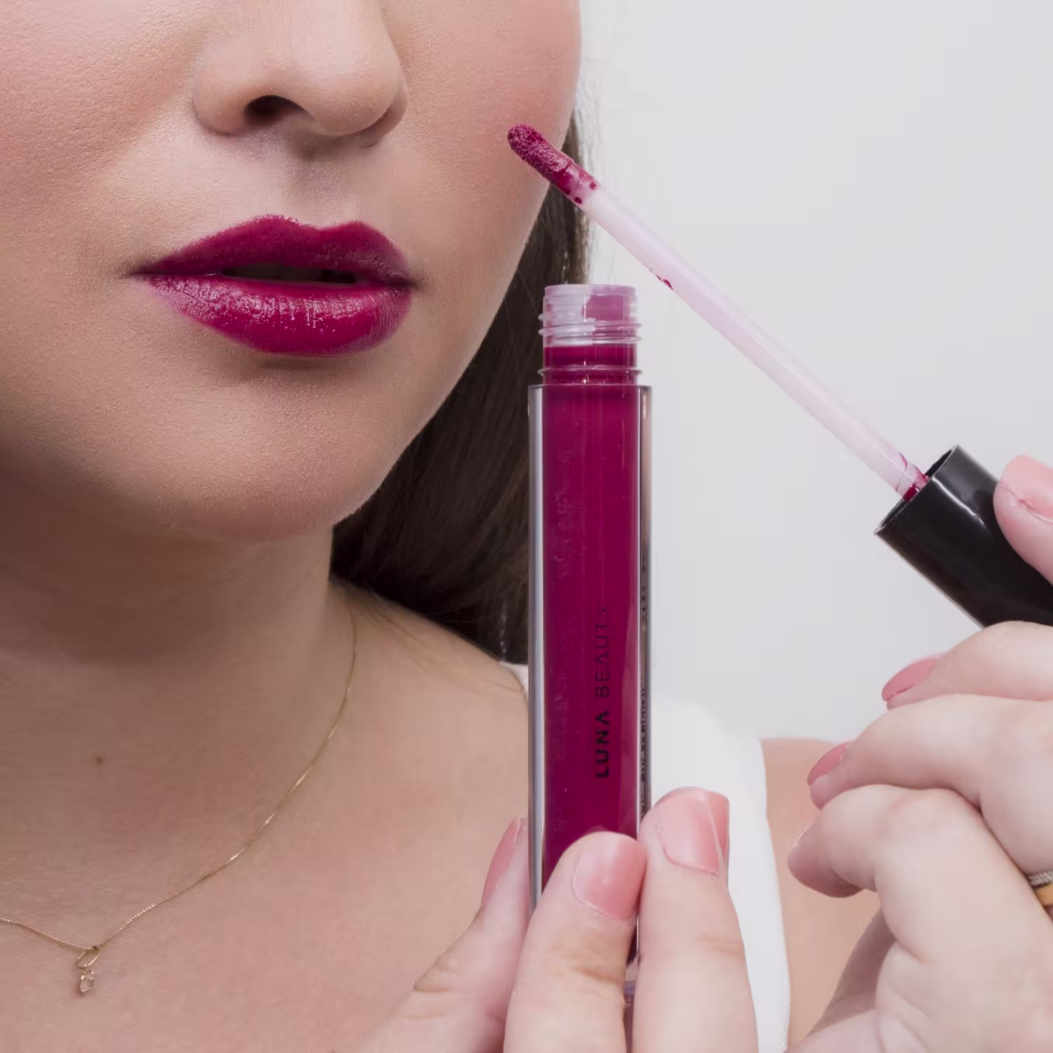 Óleo Labial em uma embalagem transparente de plástico cilíndrica. O produto tem uma cor vibrante de magenta. A embalagem mostra o nome "Luna Beauty" impresso em letras brancas. Na imagem, é visível uma mão feminina segurando o produto próximo aos lábios.