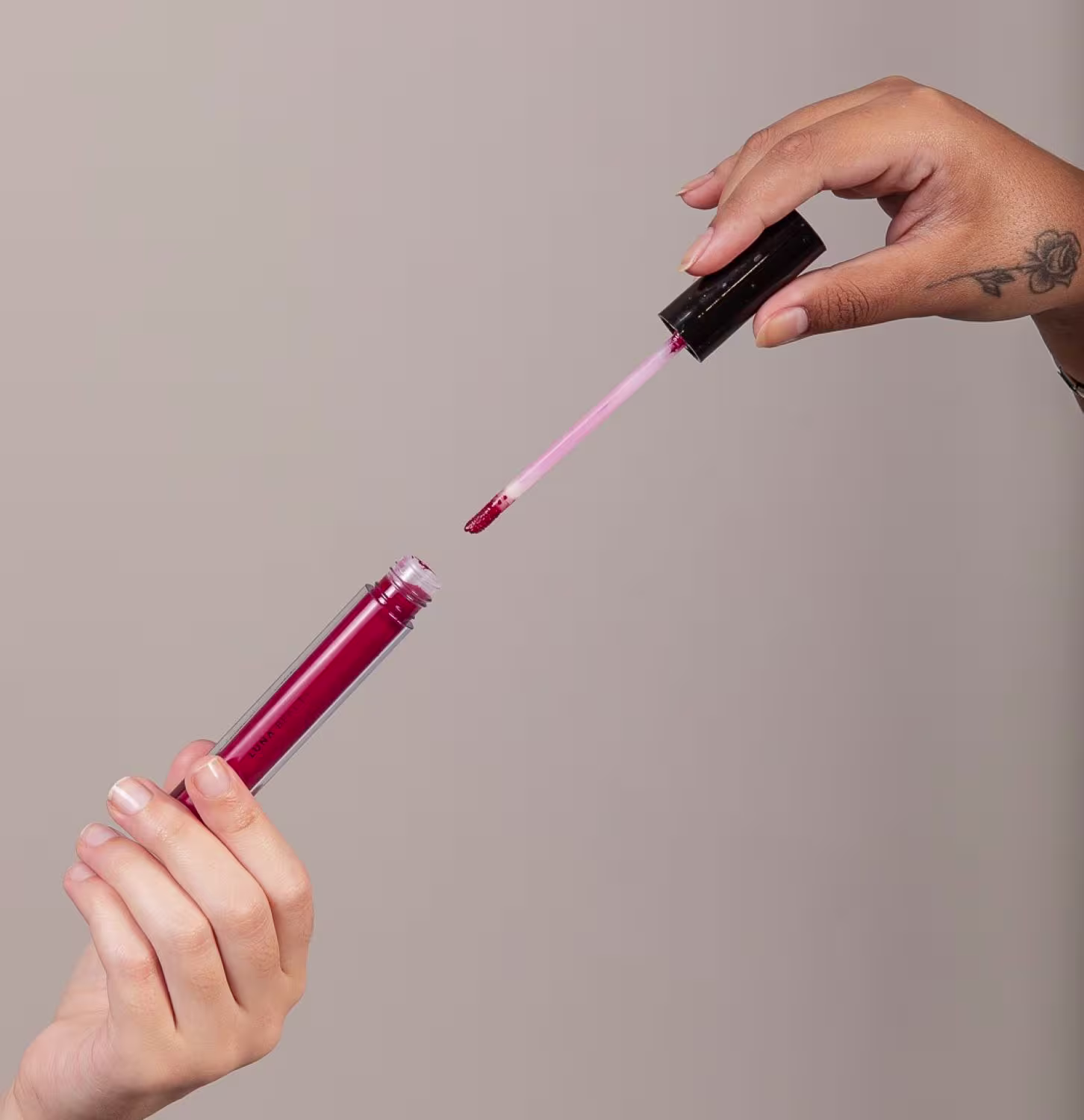 Um aplicador de batom líquido. O frasco é transparente, permitindo ver o líquido dentro. As mãos segurando o frasco e o aplicador são visíveis, uma delas apresenta uma tatuagem.