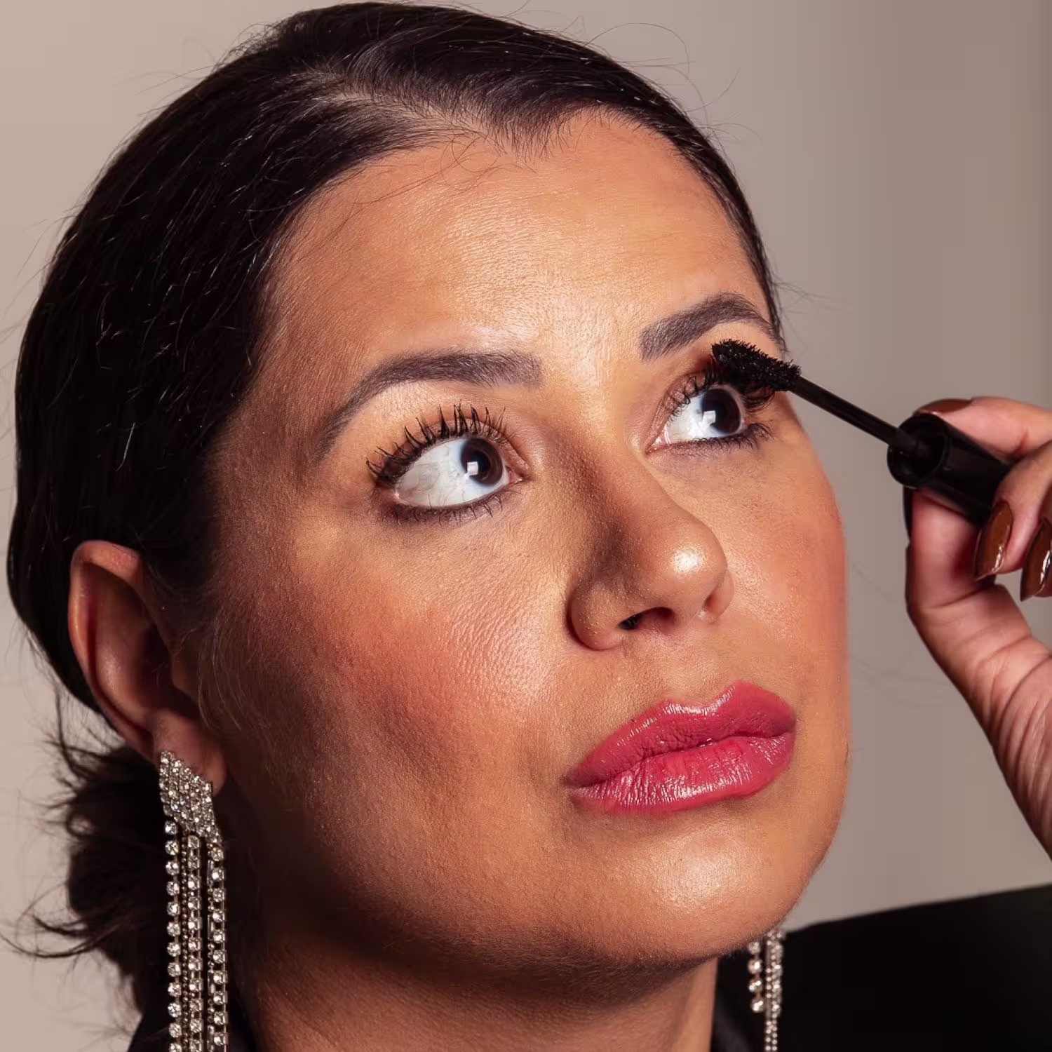 Esta imagem mostra uma mulher aplicando máscara para cílios em seus olhos. Ela tem pele bronzeada, lábios vermelhos brilhantes e está usando brincos compridos de strass.