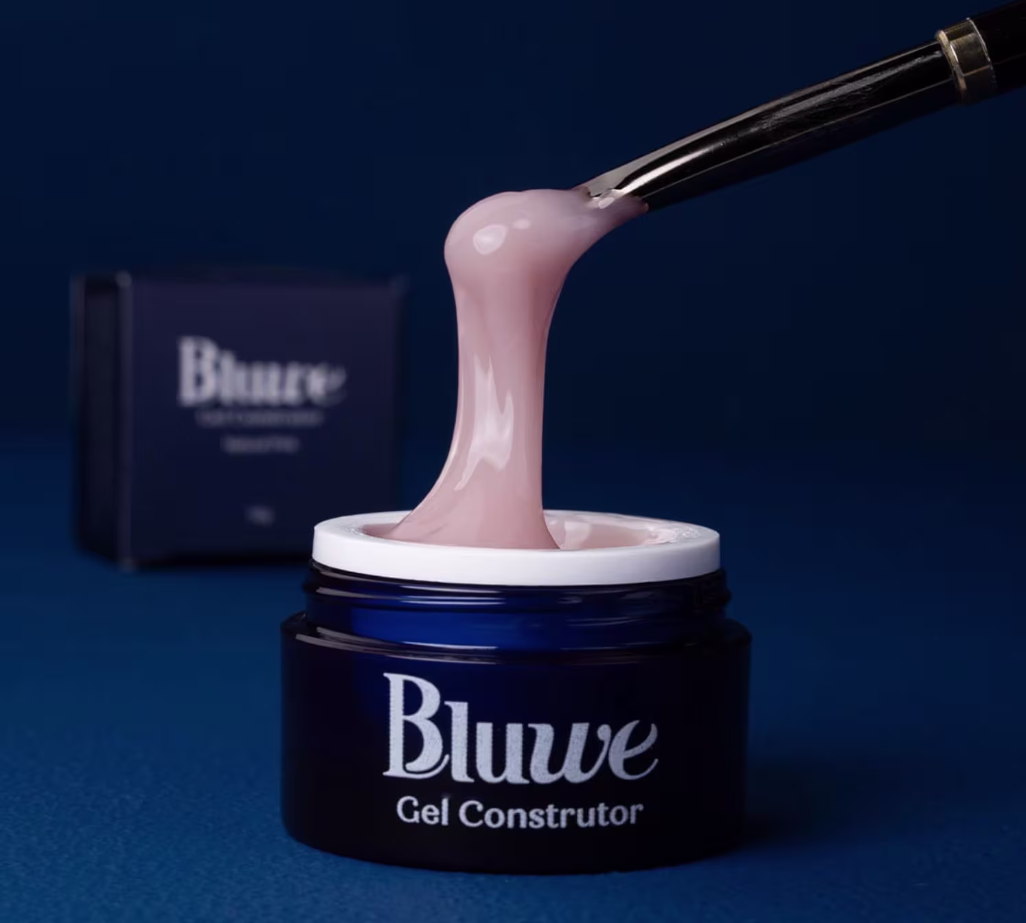 A imagem mostra o gel construtor Bluwe, em fundo azul escuro. É um frasco azul escuro com gel rosa. Há o texto "Bluwe Gel Construtor" em letras brancas e um aplicador de gel metálico longo e fino mostrando a consistência e textura do produto.
