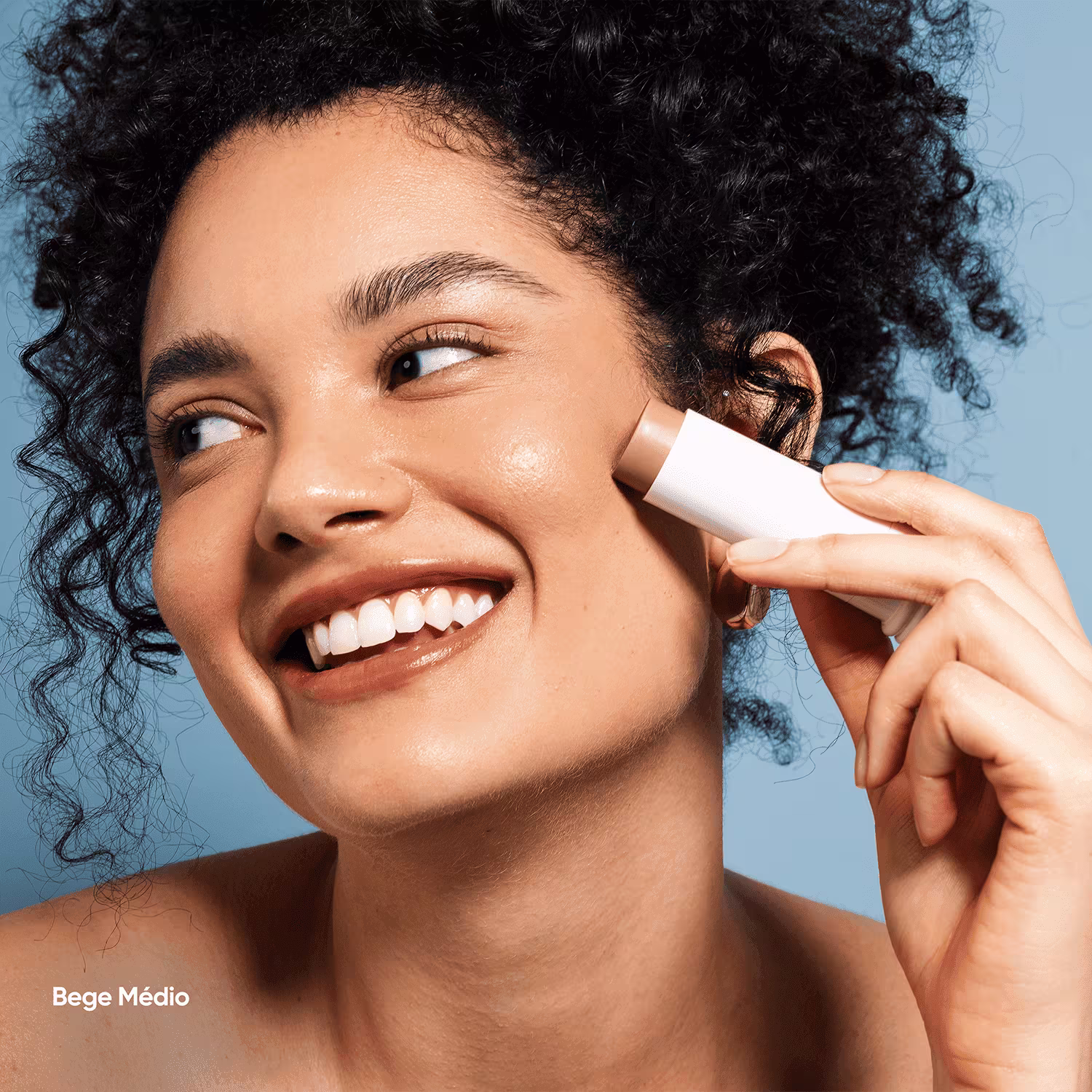 O produto apresentado é um protetor facial em formato de bastão. Possui uma embalagem cilíndrica branca. Uma mulher de pele morena clara e cabelos cacheados está sorrindo amplamente. Ela segura o protetor próximo ao rosto.