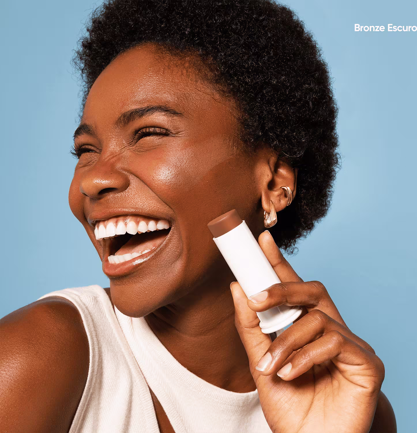 O produto apresentado é um protetor facial em formato de bastão. Possui uma embalagem cilíndrica branca. Uma mulher de pele negra e cabelos crespos está sorrindo amplamente. Ela segura o protetor próximo ao rosto.