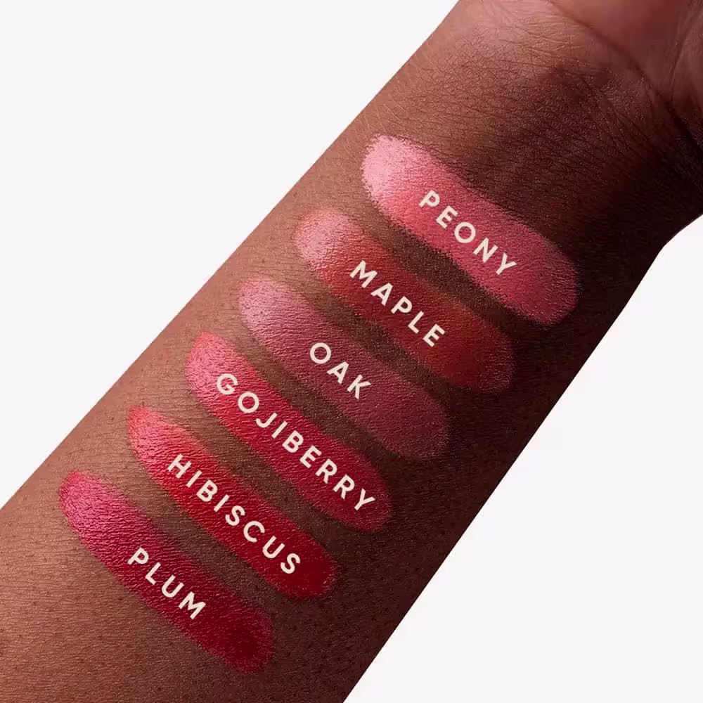 A imagem mostra uma série de amostras da cor do blush em diferentes tons de vermelho, variando de um rosa claro a um vermelho escuro. Os nomes dos tons estão impressos sobre cada amostra, incluindo "Peony", "Maple Oak", "Goji Berry" e "Hibiscus Plum".