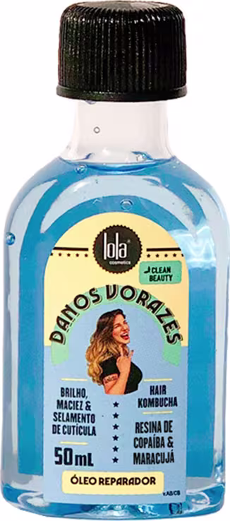 A imagem mostra o óleo capilar danos vorazes, seu frasco é pequeno transparente permitindo ver o líquido azul claro. No rótulo há uma ilustração de uma mulher. Sua tampa é preta rosqueável.