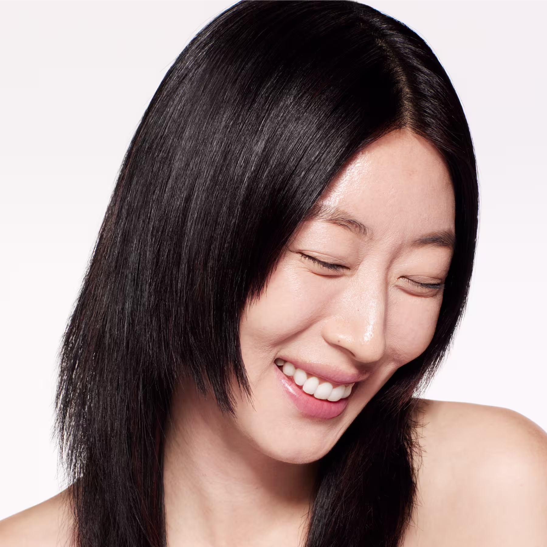 Esta imagem apresenta uma mulher asiática jovem com cabelos longos e lisos de cor preta. Ela tem uma pele clara e saudável, com um sorriso genuíno e olhos fechados. A mulher está em um estúdio com um fundo branco simples, destacando seus traços naturais.