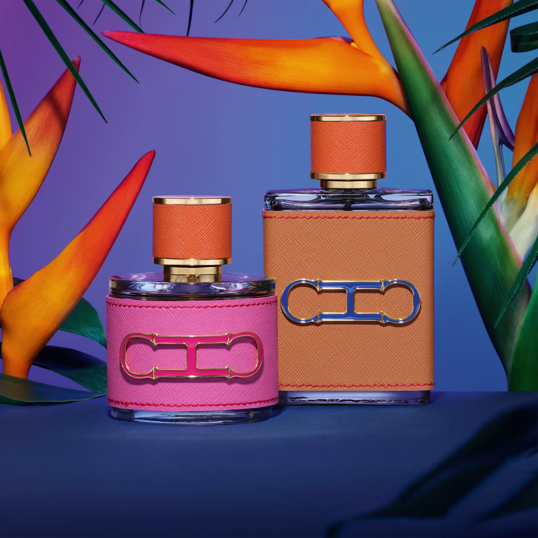 A imagem mostra dois frascos de perfumes, da mesma coleção. Um menor, arredondado, com capa rosa e tampa laranja, e um maior, retangular, com capa e tampa laranja. Ambos apresentam a logo da marca em alto relevo.