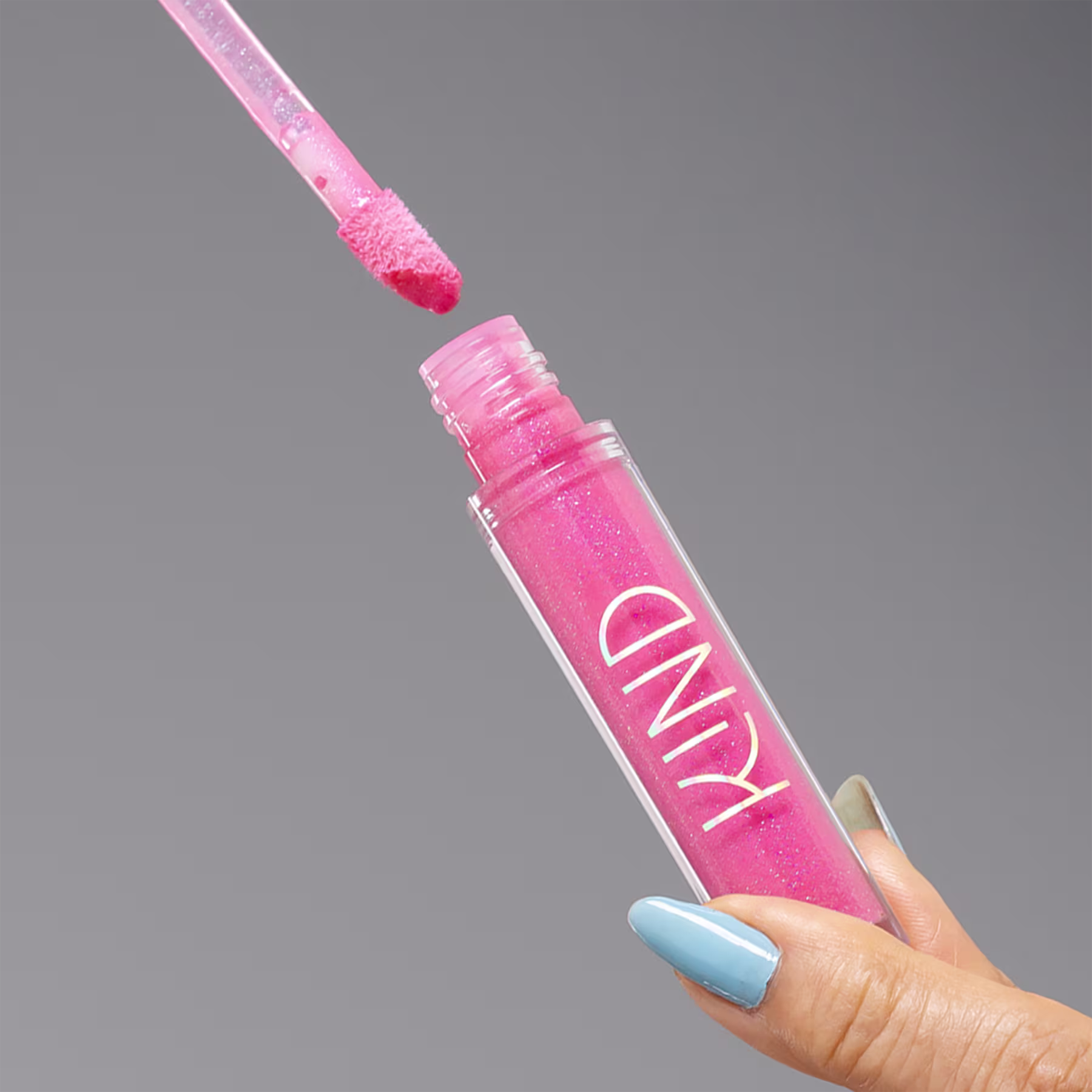 A imagem mostra a embalagem do gloss, aberta, sendo segurado por uma mão, com unhas azuis.