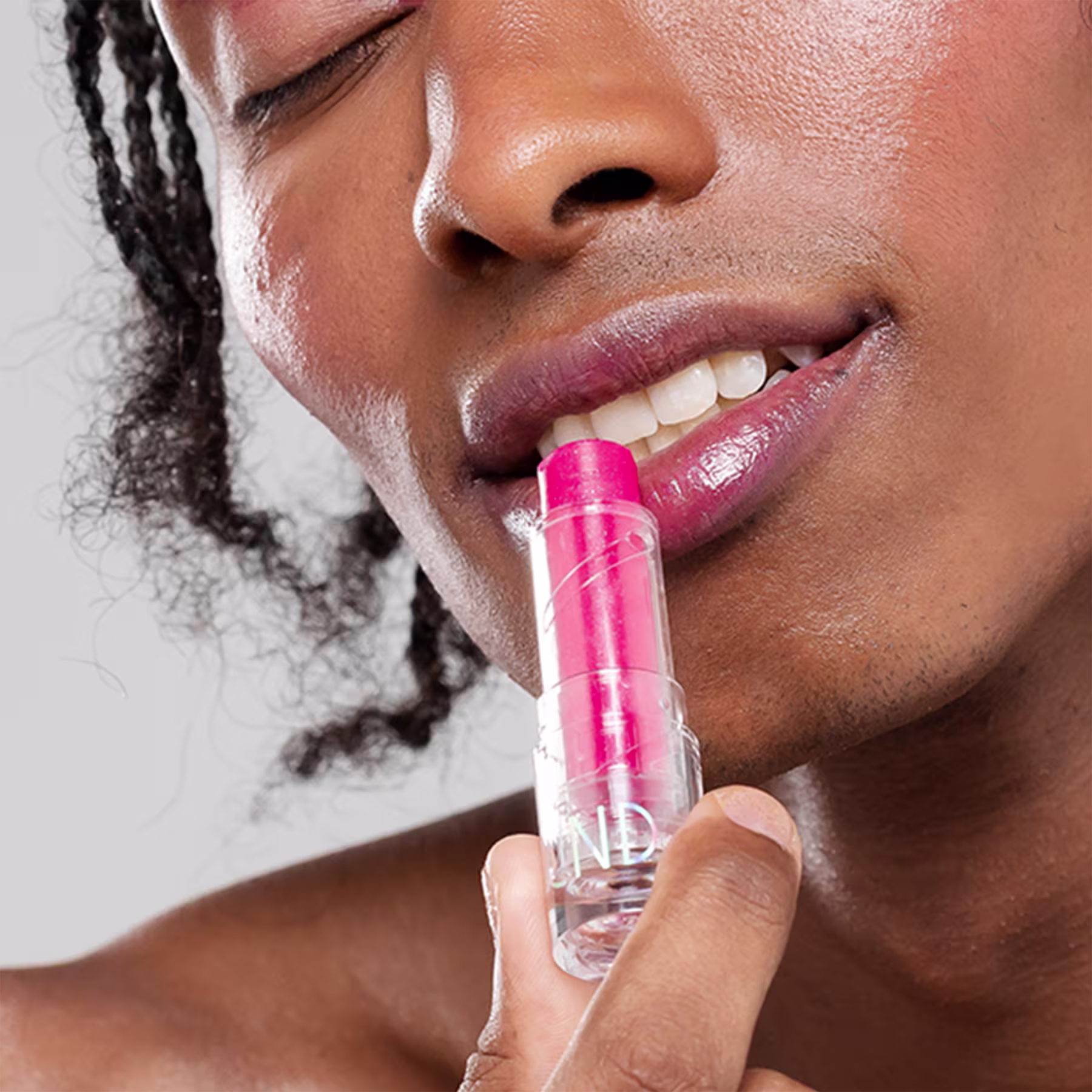 A imagem mostra o rosto de uma pessoa negra, usando o balm e segurando a embalagem, com uma das mãos.