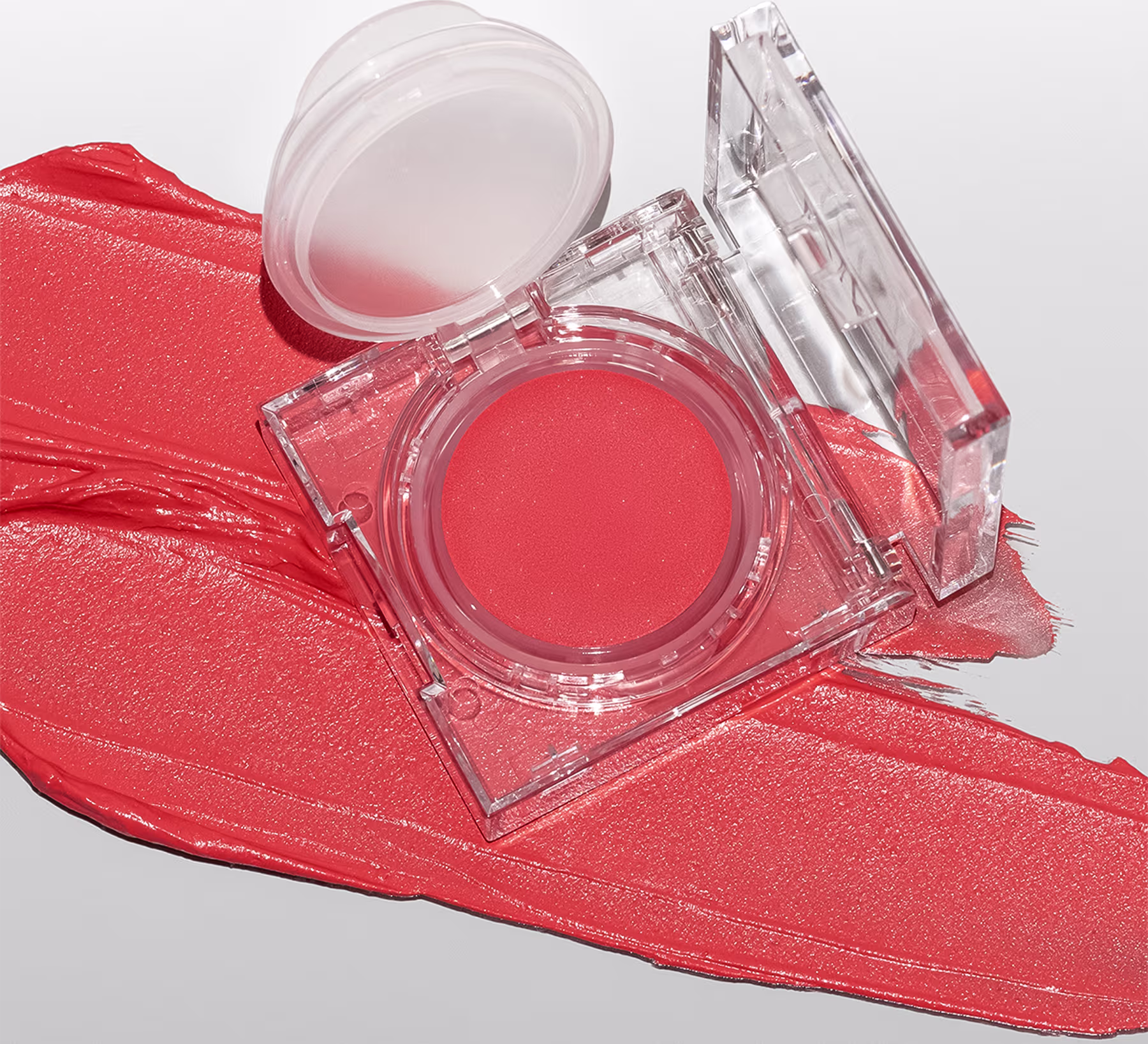 A imagem mostra a embalagem do blush cremoso aberta, numa superfície branca com um pouco do blush esparramado pela superfície, mostrando a cor e a textura cremosa.