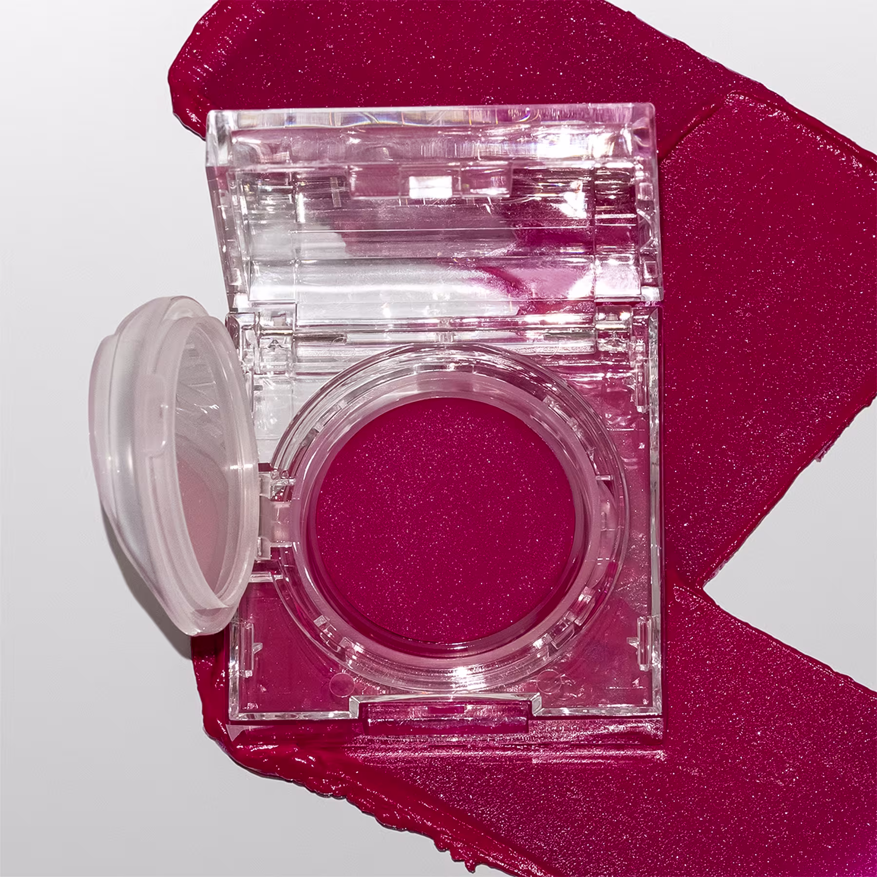 A imagem mostra a embalagem do blush cremoso aberta, numa superfície branca com um pouco do blush esparramado pela superfície, mostrando a cor e a textura cremosa.