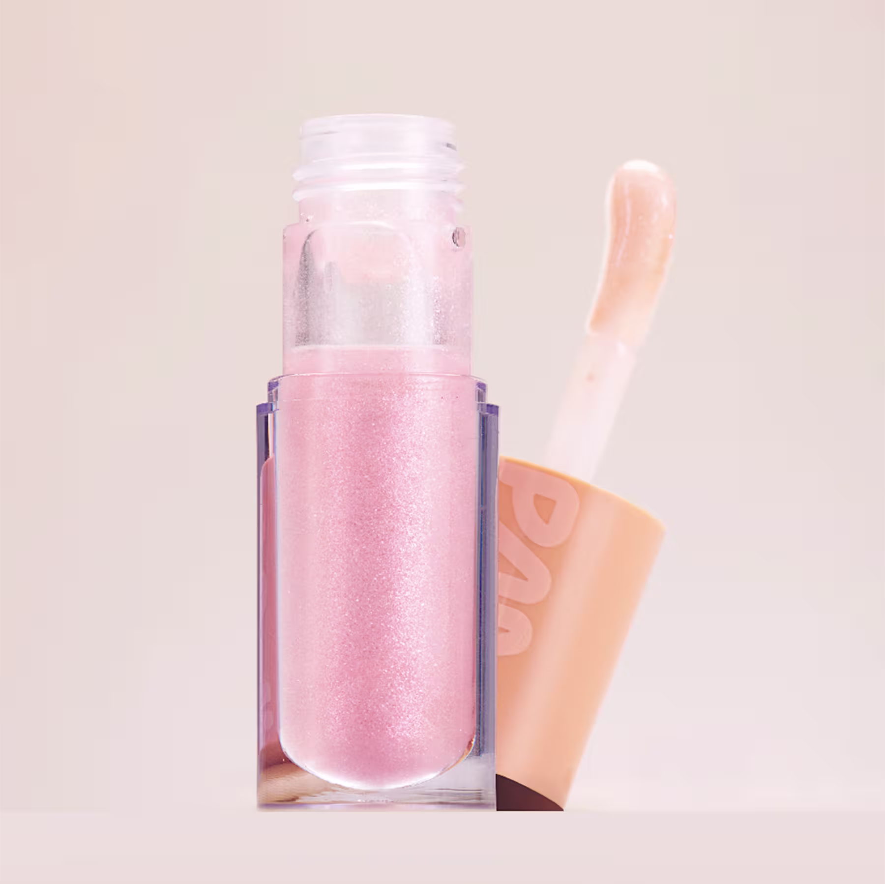 A imagem mostra o gloss labial da marca Pam! By Pamella. Seu tubo é transparente e permite ver sua cor rosa claro vibrante. Ao lado tem seu aplicador na mesma tonalidade rosa claro. Ambos ostentam o logotipo PAM estampado na tampa laranja.