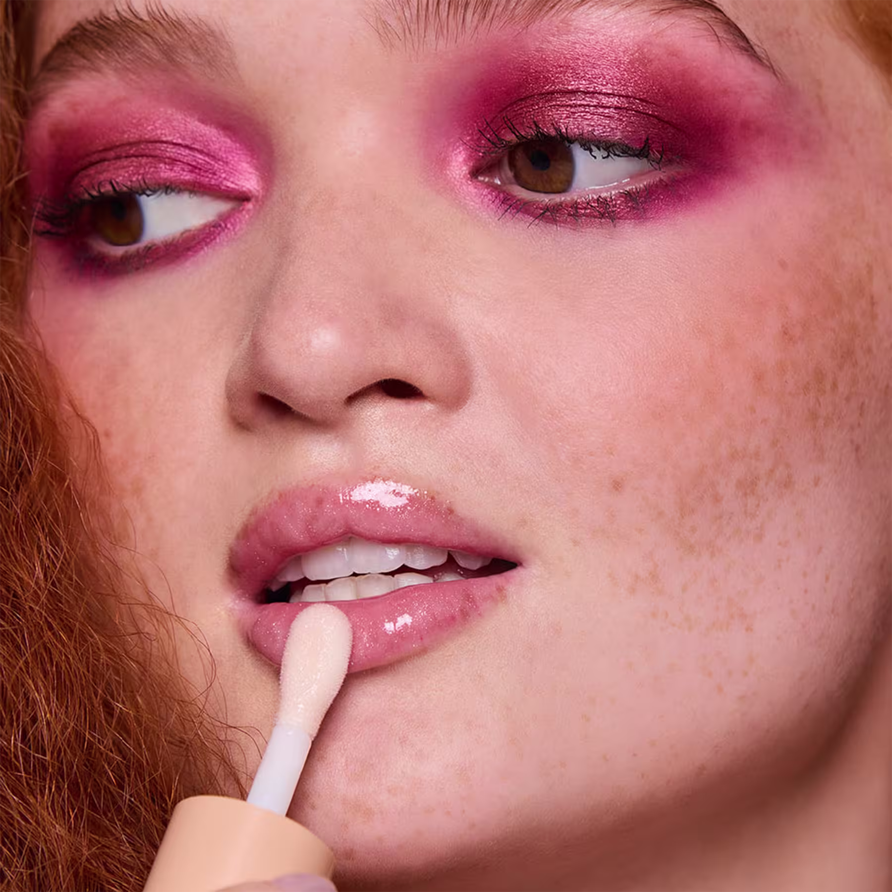 Esta imagem mostra uma mulher ruiva com maquiagem rosa vibrante potente e marcante nos olhos e nos lábios ela está aplicando o gloss labial rosa claro.