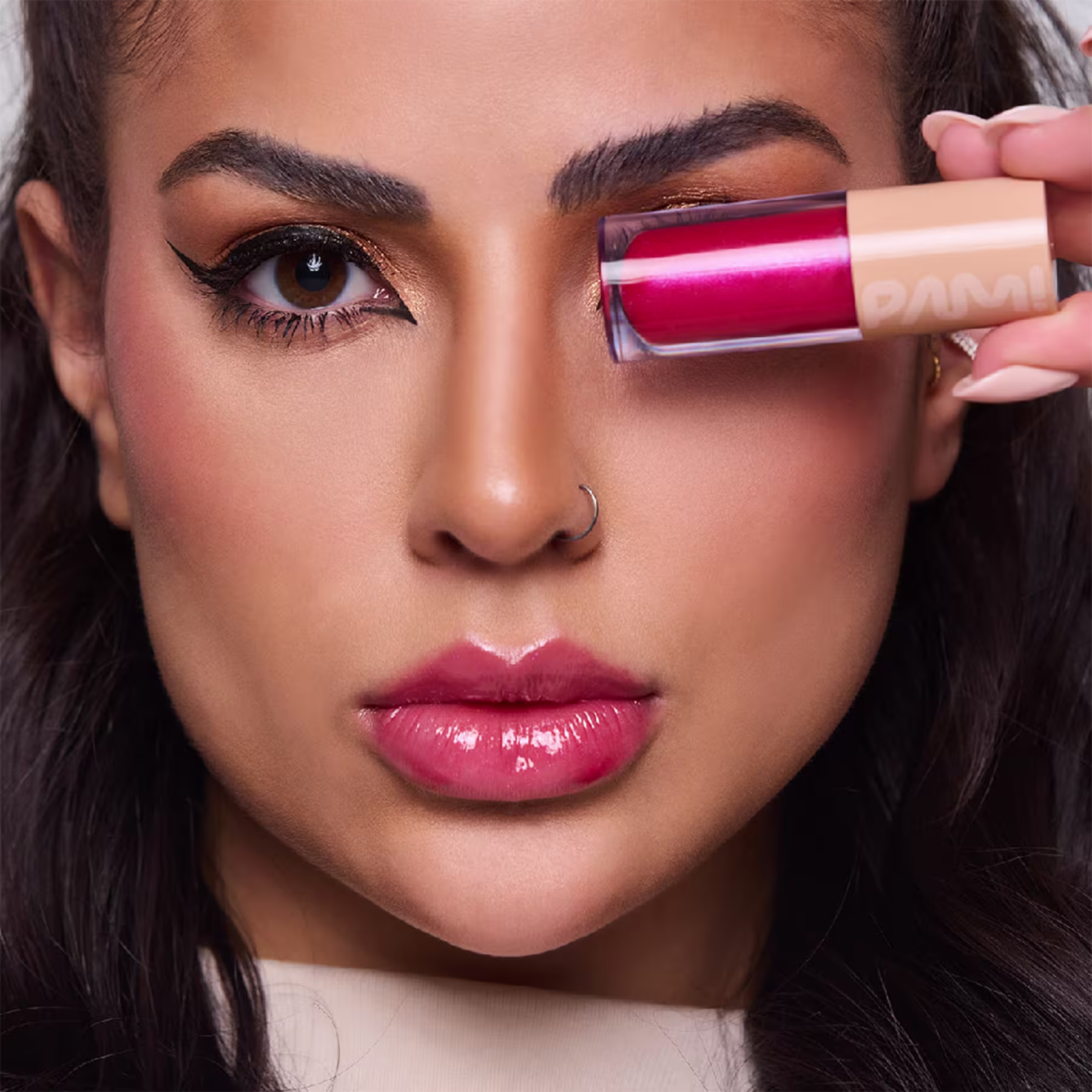 A imagem mostra um close-up de uma mulher branca de pele bronzeada, olhos marcantes com maquiagem bem delineadas. Seus lábios estão cobertos por um tom rosado brilhante. Na mão, ela segura o tubo do gloss labial de mesma cor.