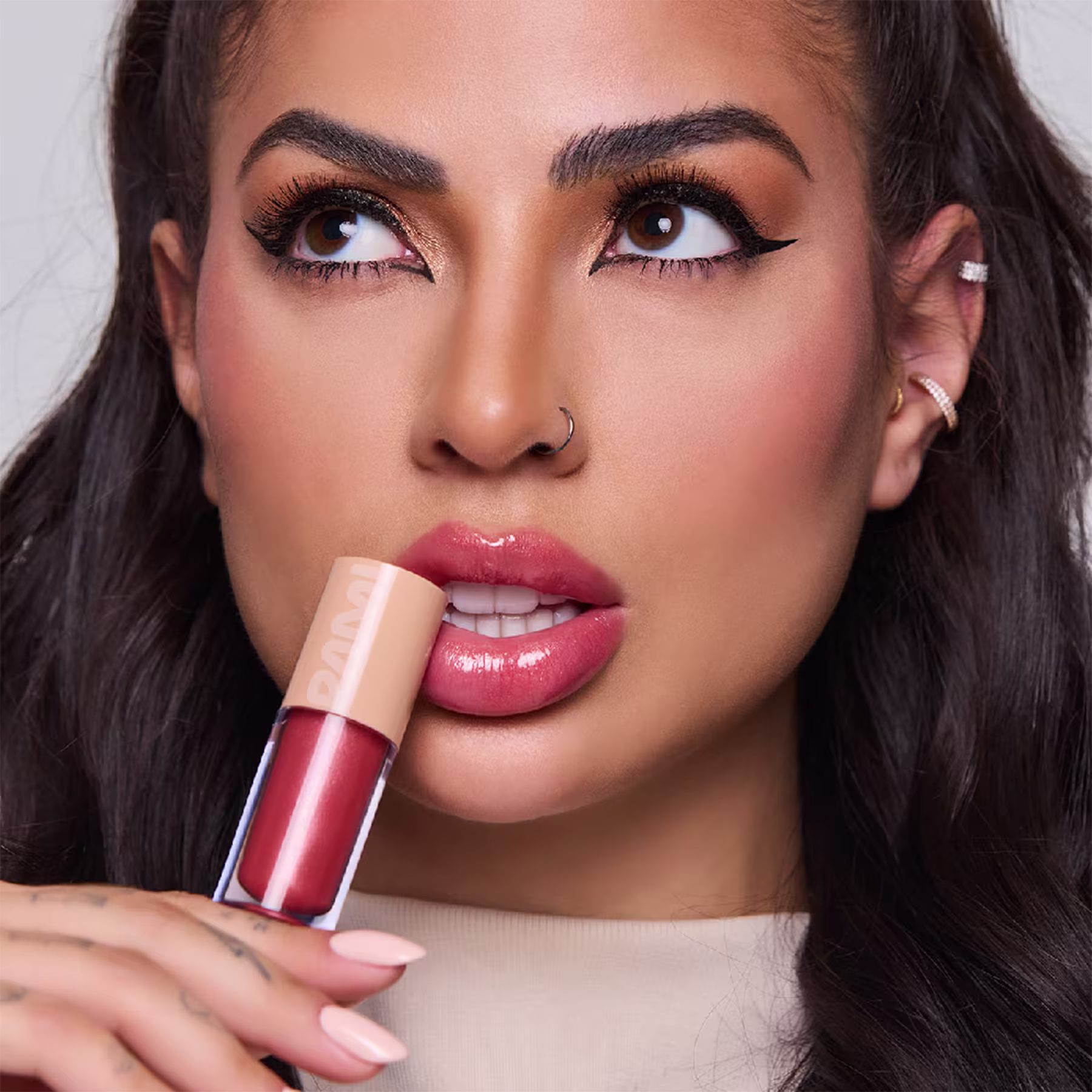 A imagem mostra uma bela mulher segurando o brilho labial, apoiando-o em seus lábios, que estão com a mesma cor rosada vibrante. Ela está exibindo uma maquiagem impecável.