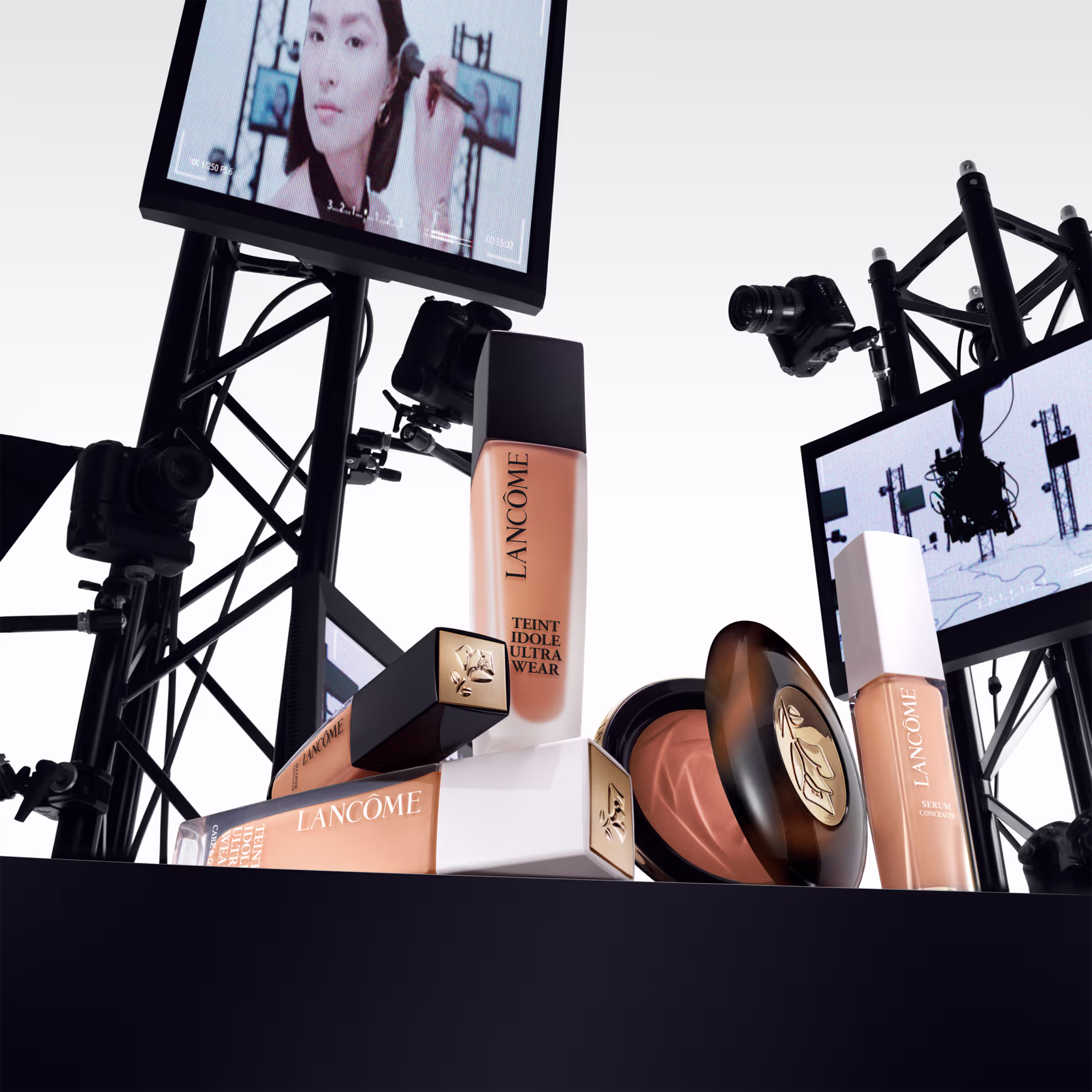 A imagem mostra alguns produtos de maquiagem Lancôme. Frascos de base líquida, na cor bege, embalagens cilíndricas com tampas pretas e brancas, com rótulos pretos e o logotipo da marca. Ao fundo alguns monitores como em um estúdio de gravação.