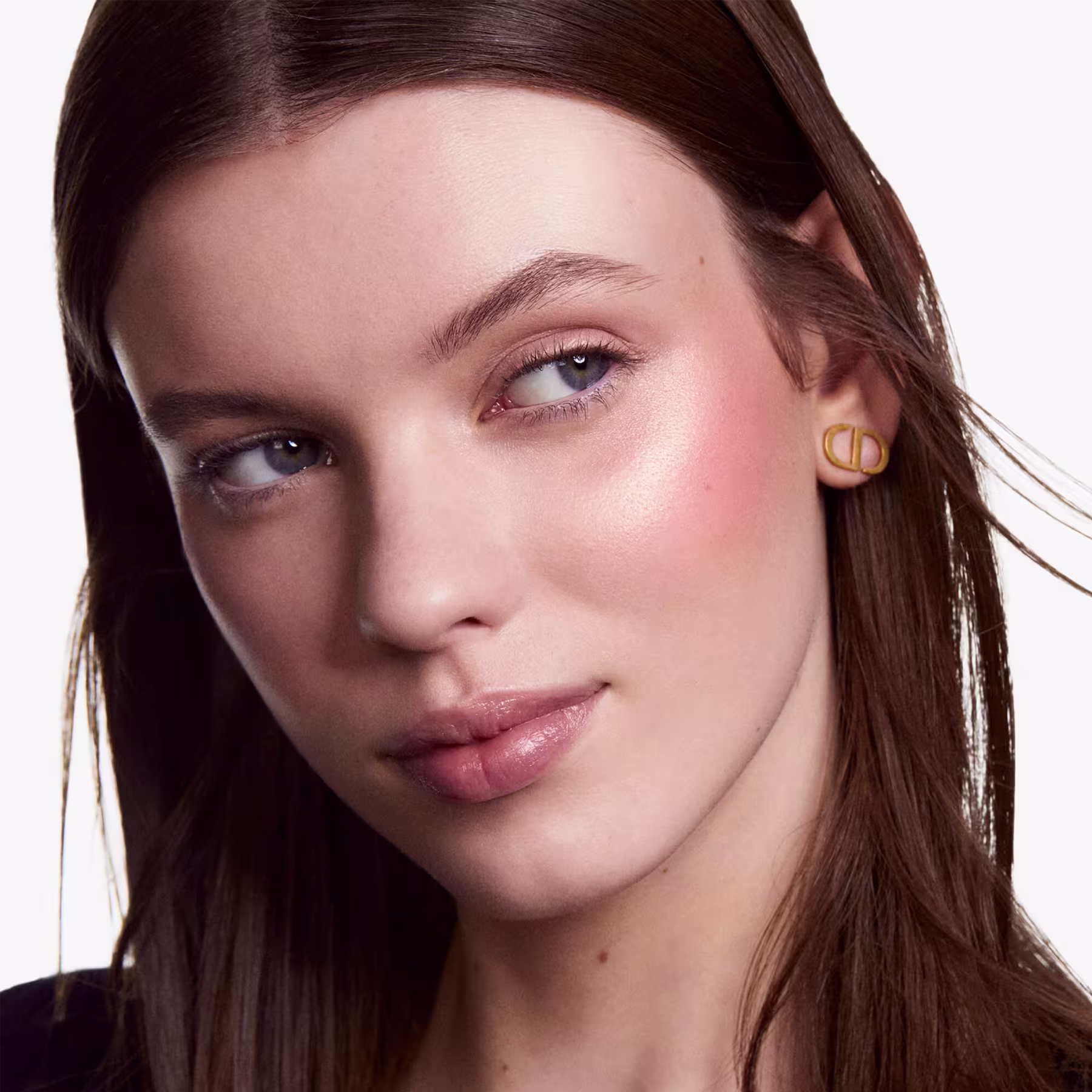 A imagem mostra uma mulher jovem com cabelos castanhos escuros e pele clara. Ela está usando maquiagem natural, com sombra marrom suave nos olhos, delineador preto discreto e batom rosa claro brilhante nos lábios cheios.