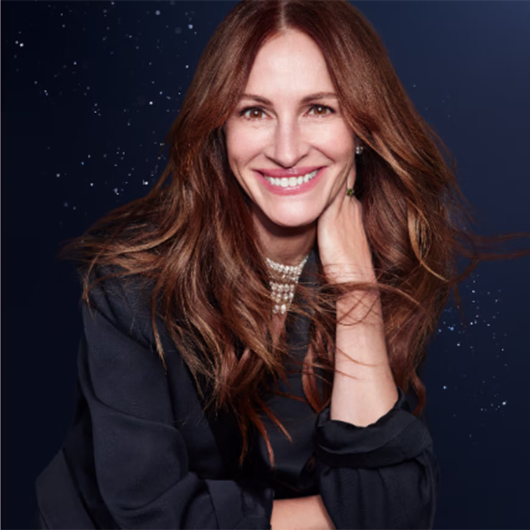 Esta imagem não mostra um produto de beleza da Lancôme. Em vez disso, é um retrato de uma mulher sorridente com longos cabelos castanhos ondulados. Ela está usando um suéter preto e um colar de pérolas.