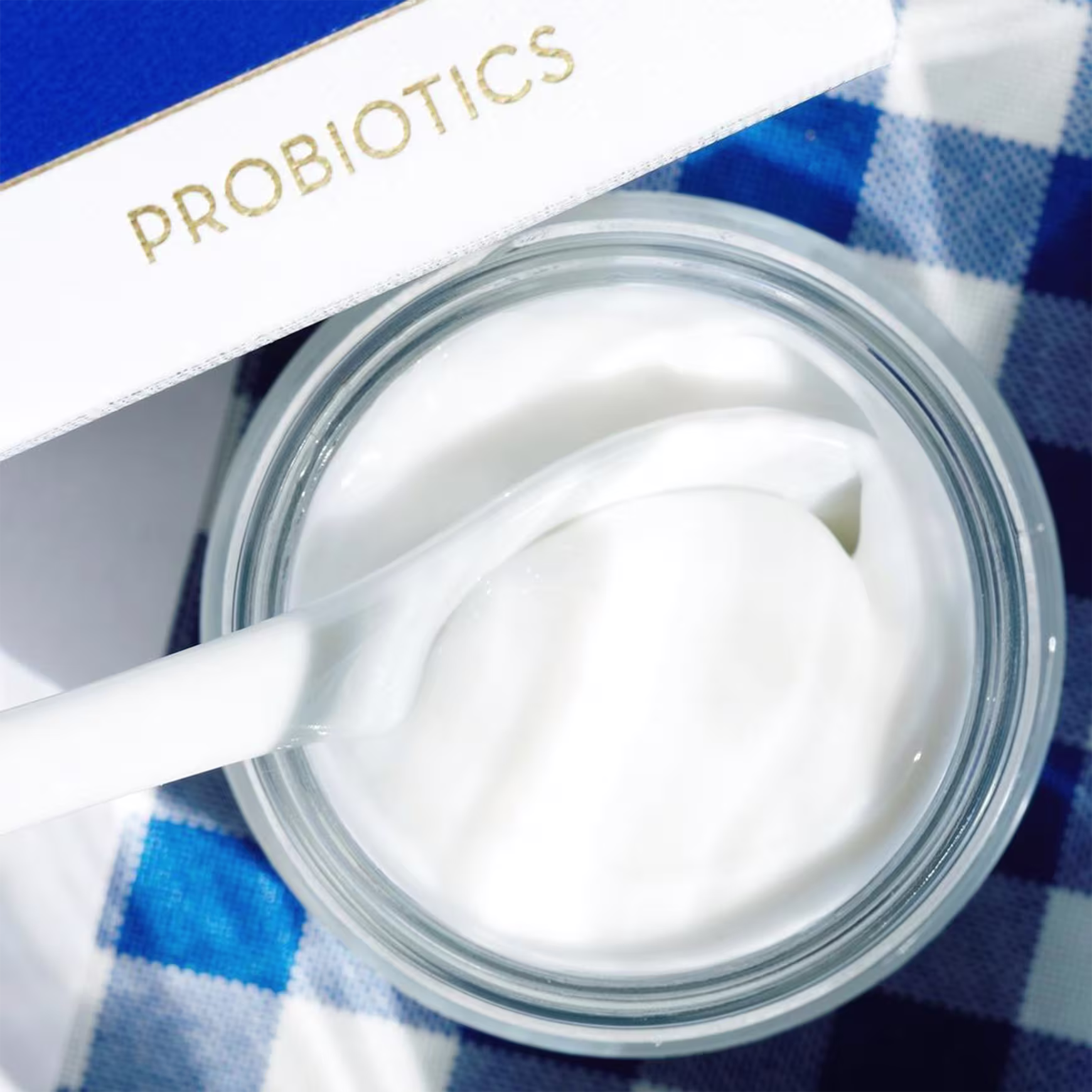 A imagem mostra a embalagem transparente aberta e uma colher branca dentro do produto. O produto está posicionado sobre um tecido listrado azul e branco.